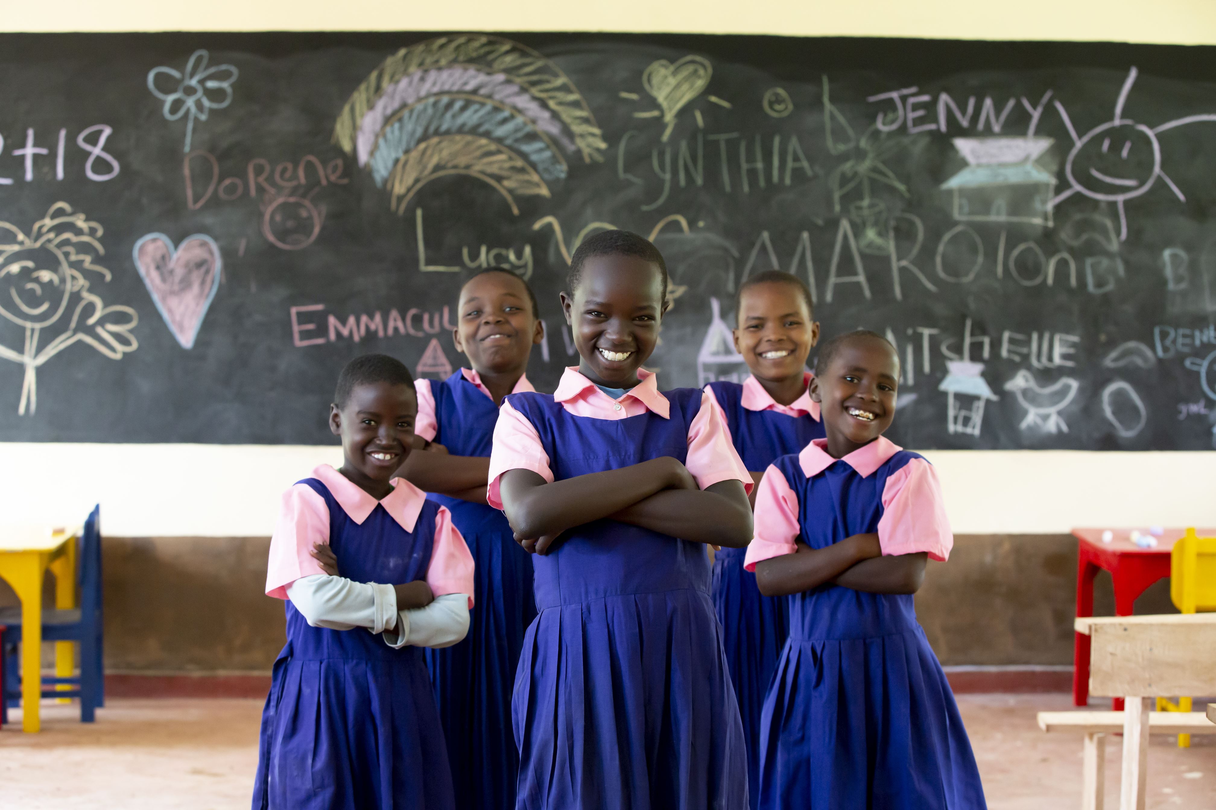 Kenyan school girls, in pink and blue uniform, stand together in front of their classroom blackboard
