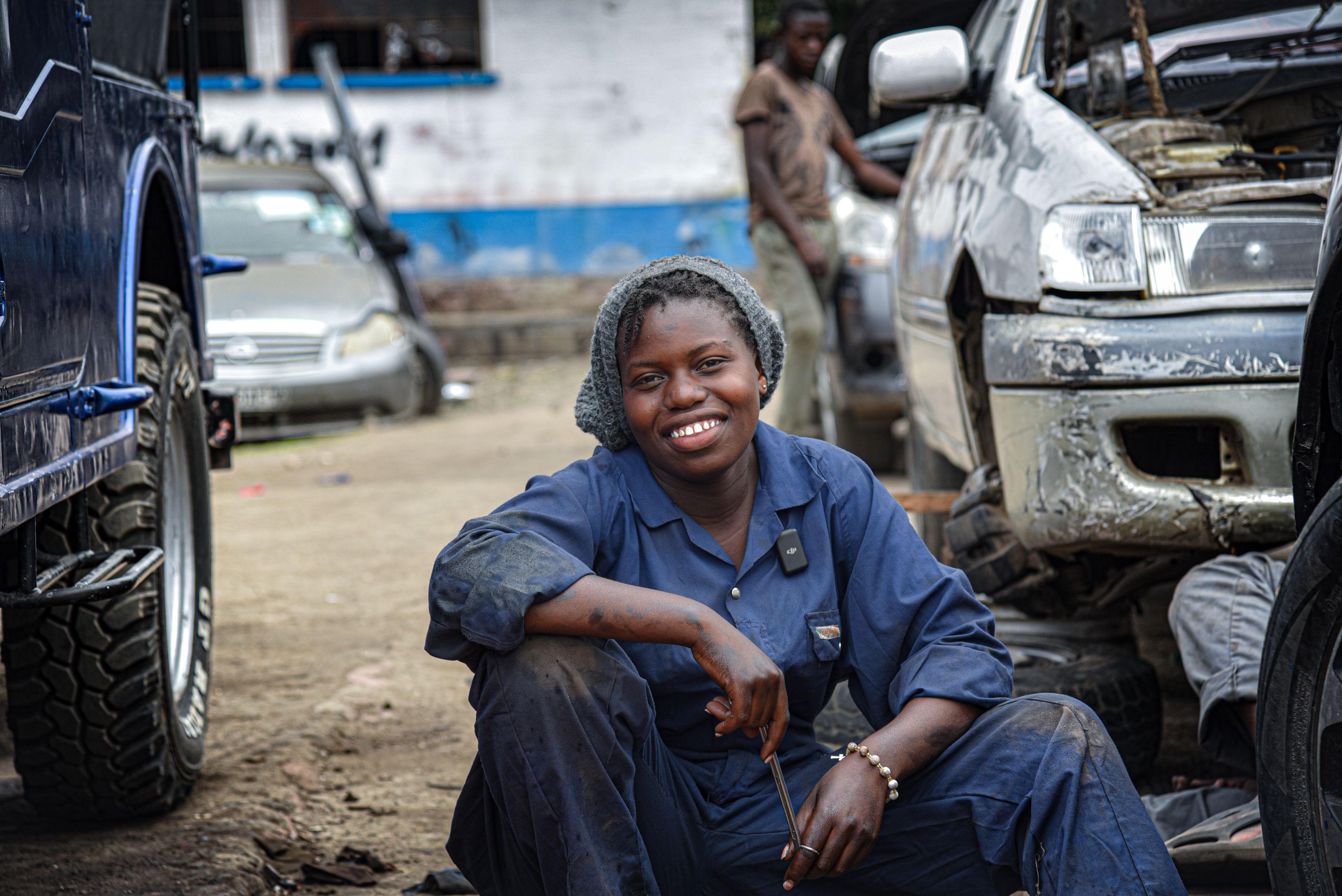 18-year-old Immaculée smiles, sitting between cars in her dark blue mechanic overalls