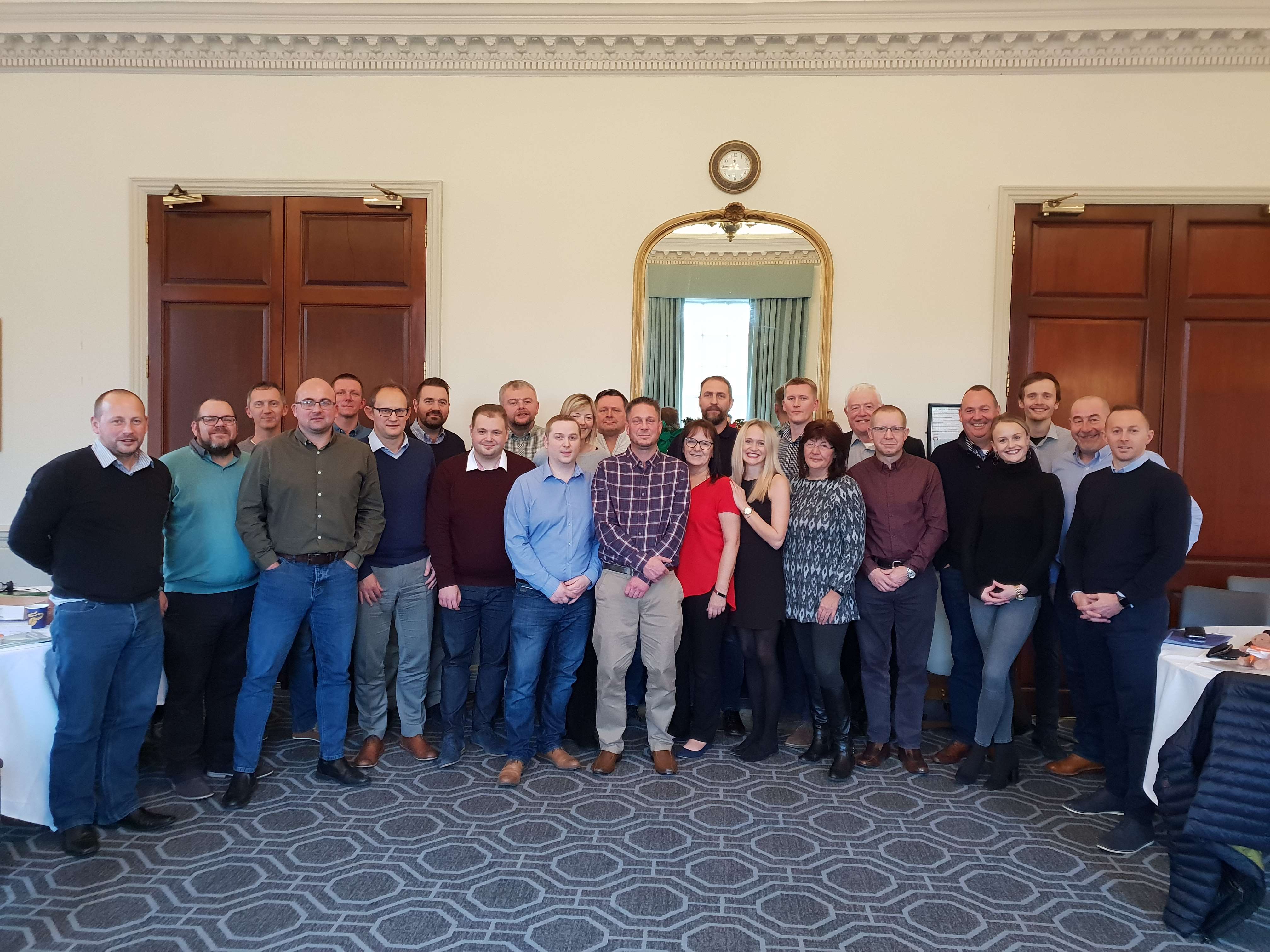 A group of around 20 people in the UK stand in a room looking at a camera