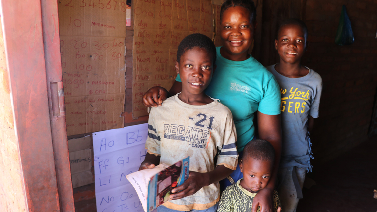 Zambian boy stands in the reading corner in his home, with his mum and siblings