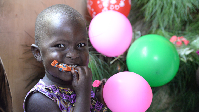 Child chews on a sweet whilst looking at the camera and sitting in front of pink and green balloons