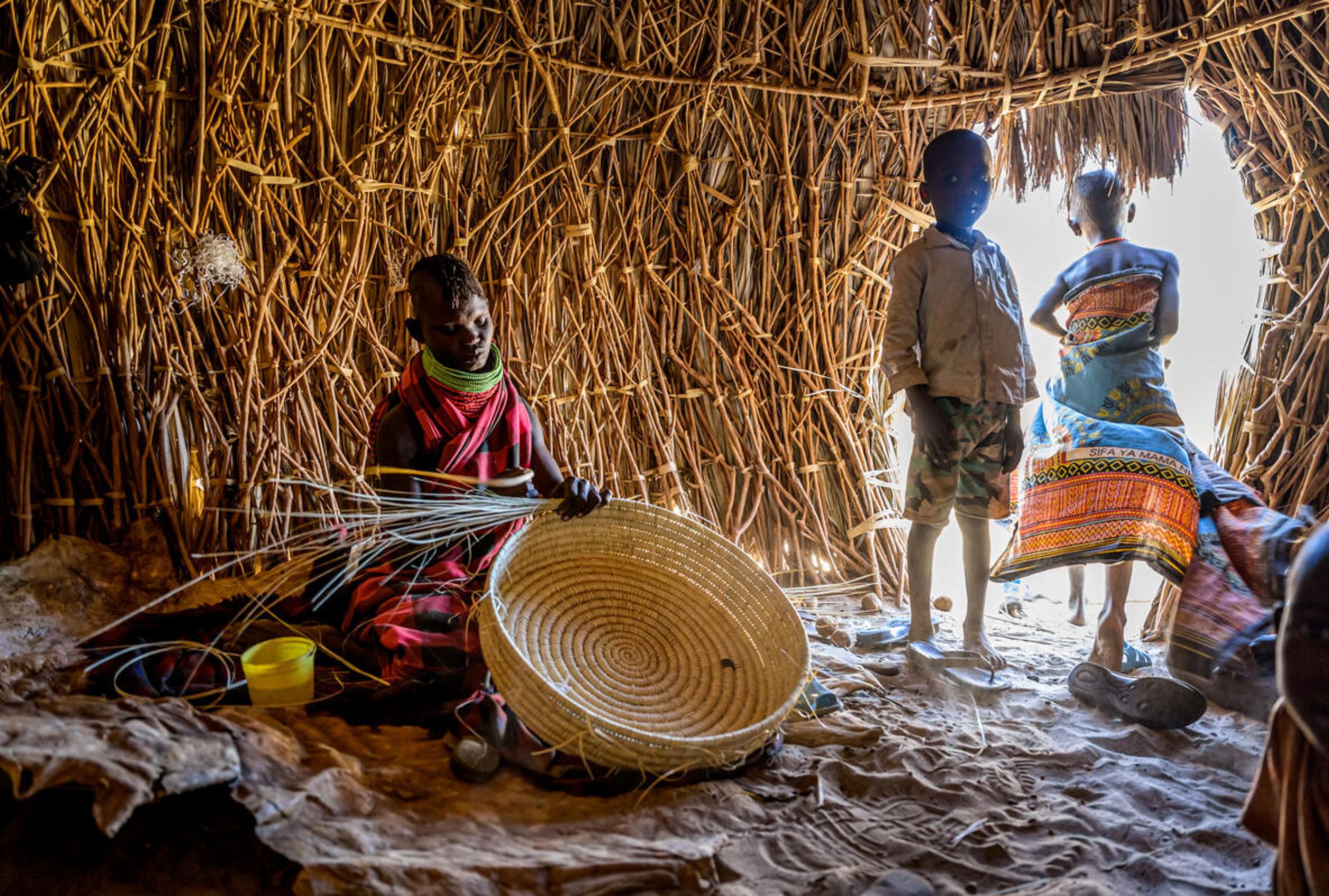 Kenya mother weaving a basket inside her straw home next to her children