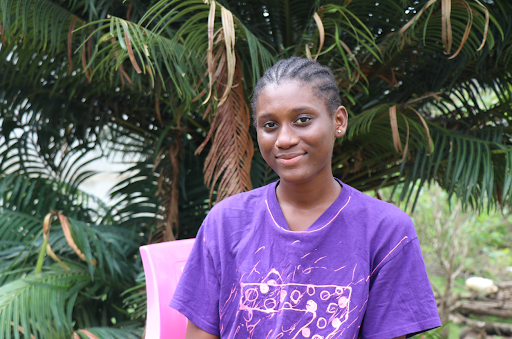 A teenage girl with braided hair sits in front of trees in Sierra Leone