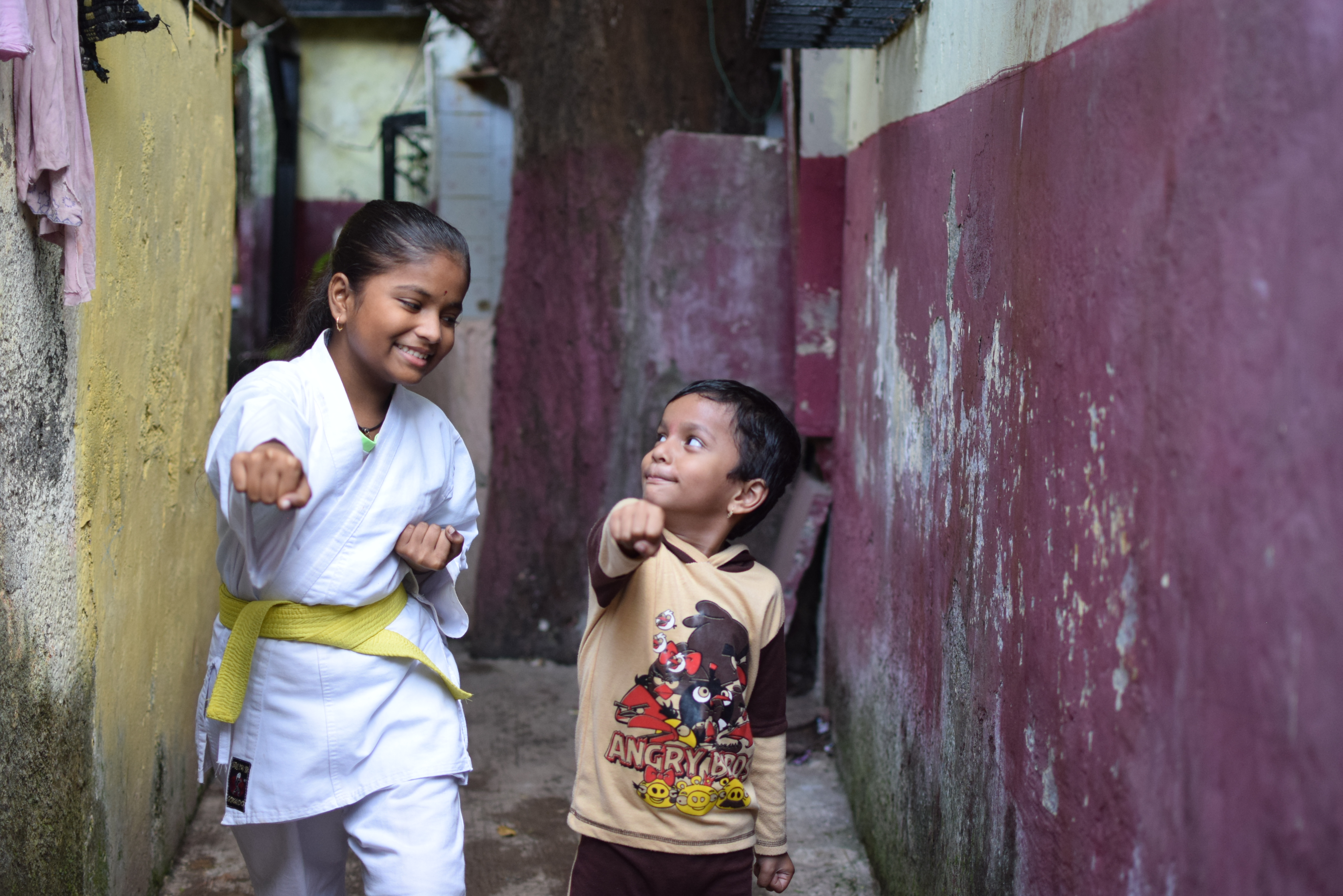 14-year-old girl in karate kit with yellow belt, holds a karate pose. Her little brother stands next to her, copying