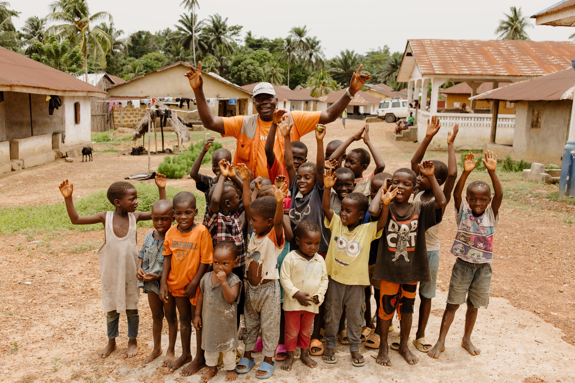 A World Vision staff member in Sierra Leone stands surrounding by children. All are lifting their hands in the air as they smile to camera.