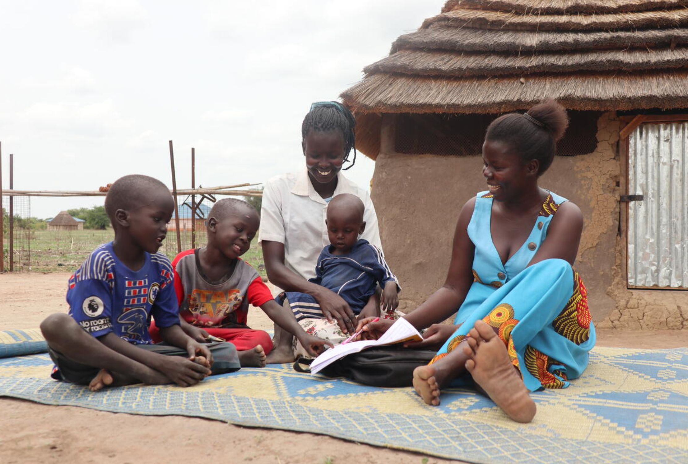 Two South Sudanese women and three children sit on the floor outside their home