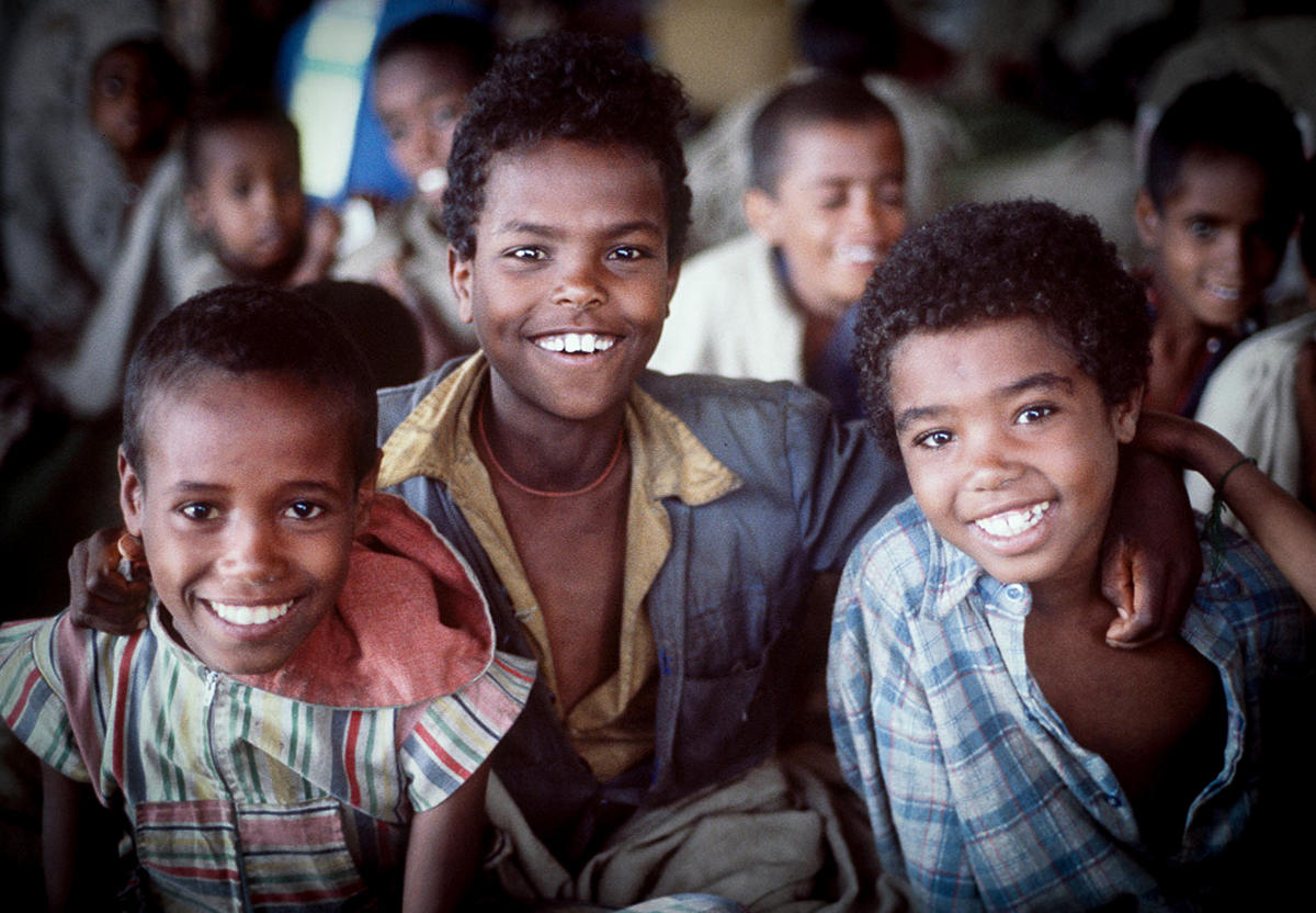 Three smiling Ethiopian children sit together in a camp in 1985