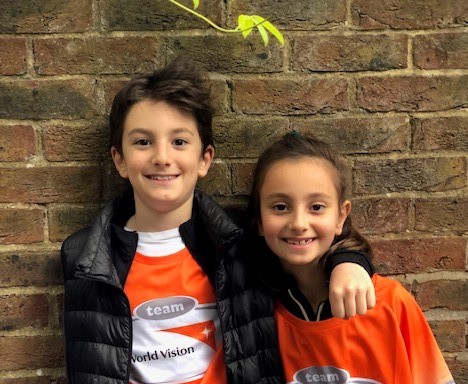 Two children in the UK wear Global 6K tshirts, and stand in front of a wall, the boy with his arm around the girl