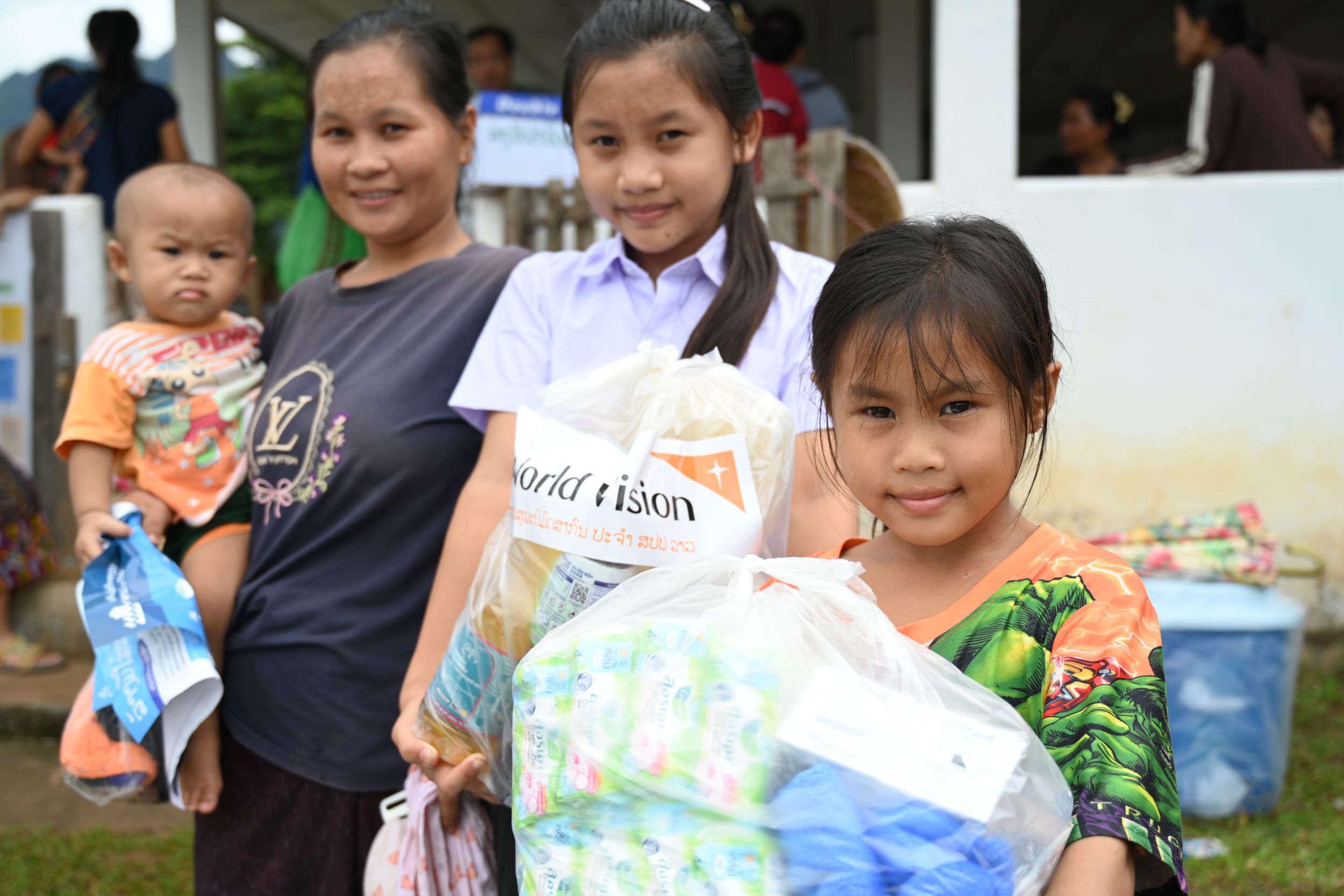 A woman from Laos holds her baby and smiles along with her two daughters, who hold food and hygiene items