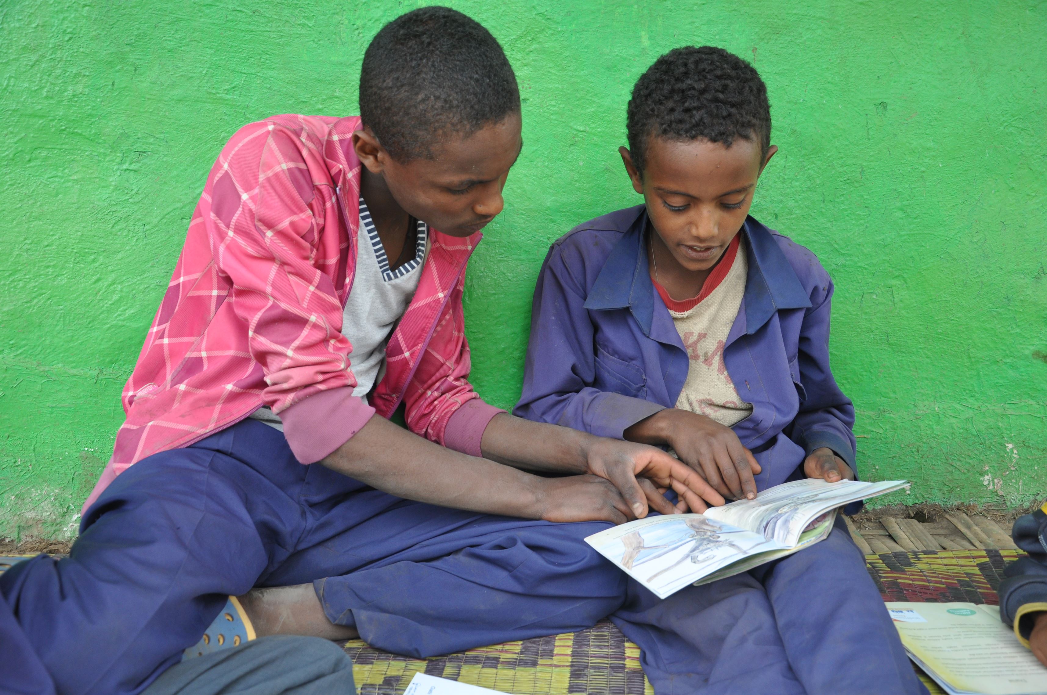 Adult in Ethiopia leans over as they help a child holding a book learn to read