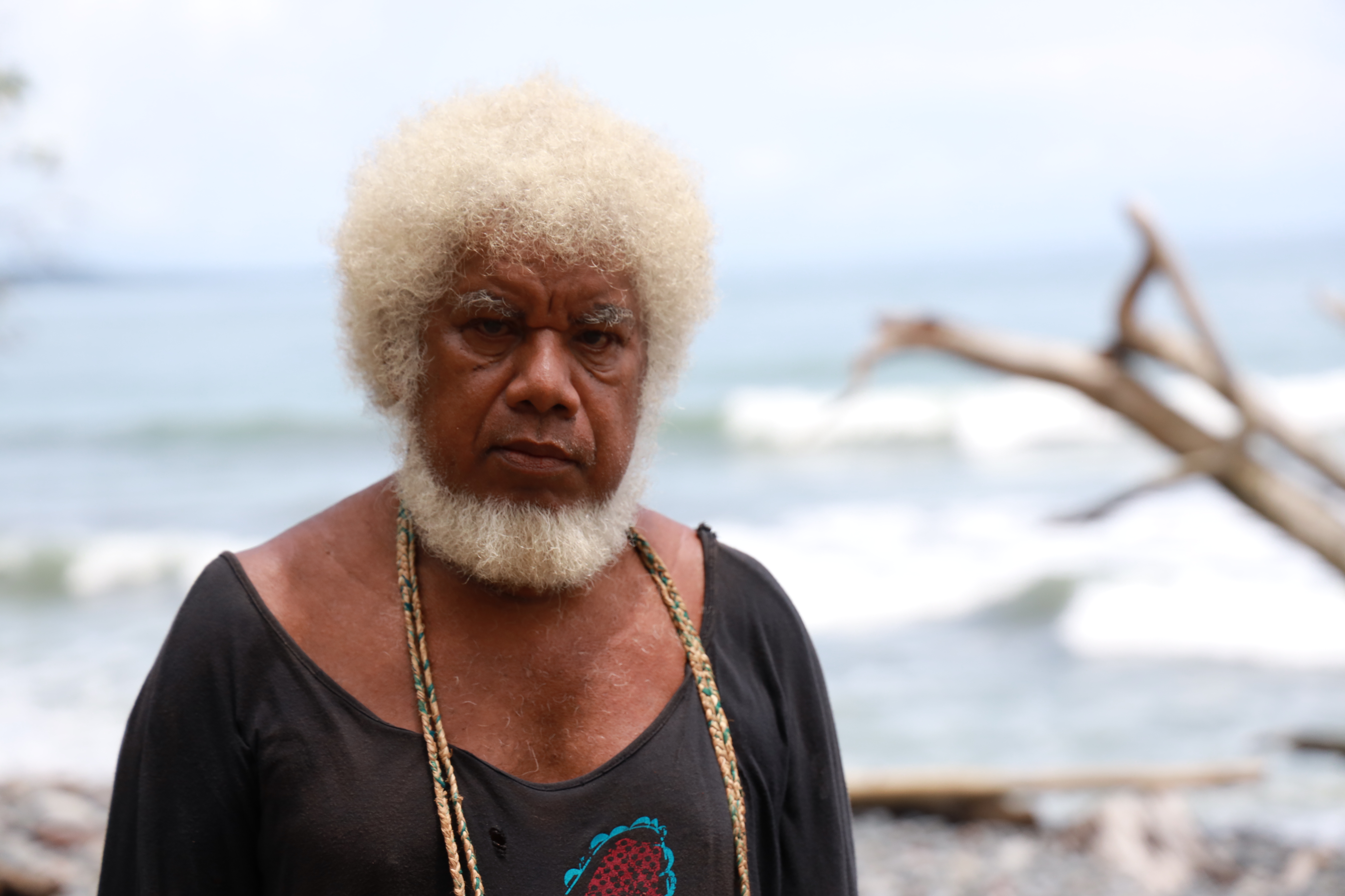 An older man stands with the sea behind him