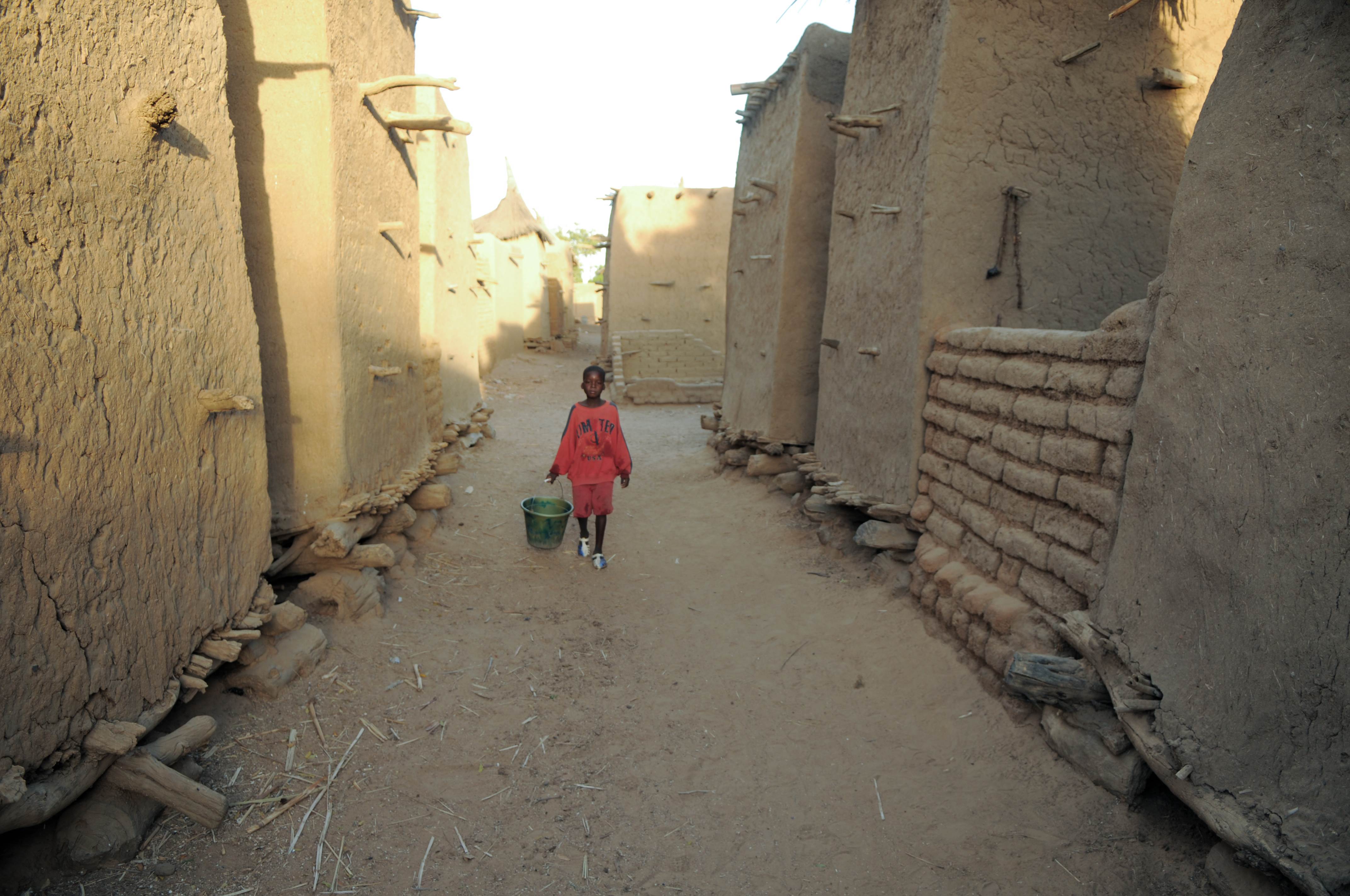 Wide shot between buildings in a village in Mali. A young boy walks with a bucket to collect water
