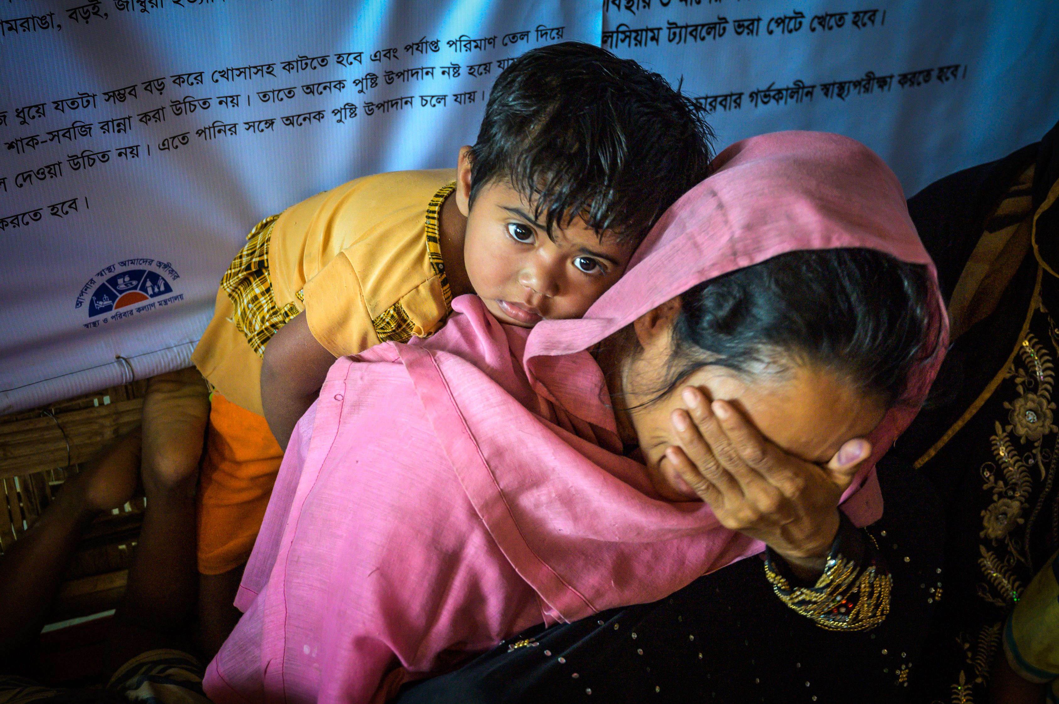 A refugee woman in pink holds a palm over her face, as she carries a child on her back in Bangladesh