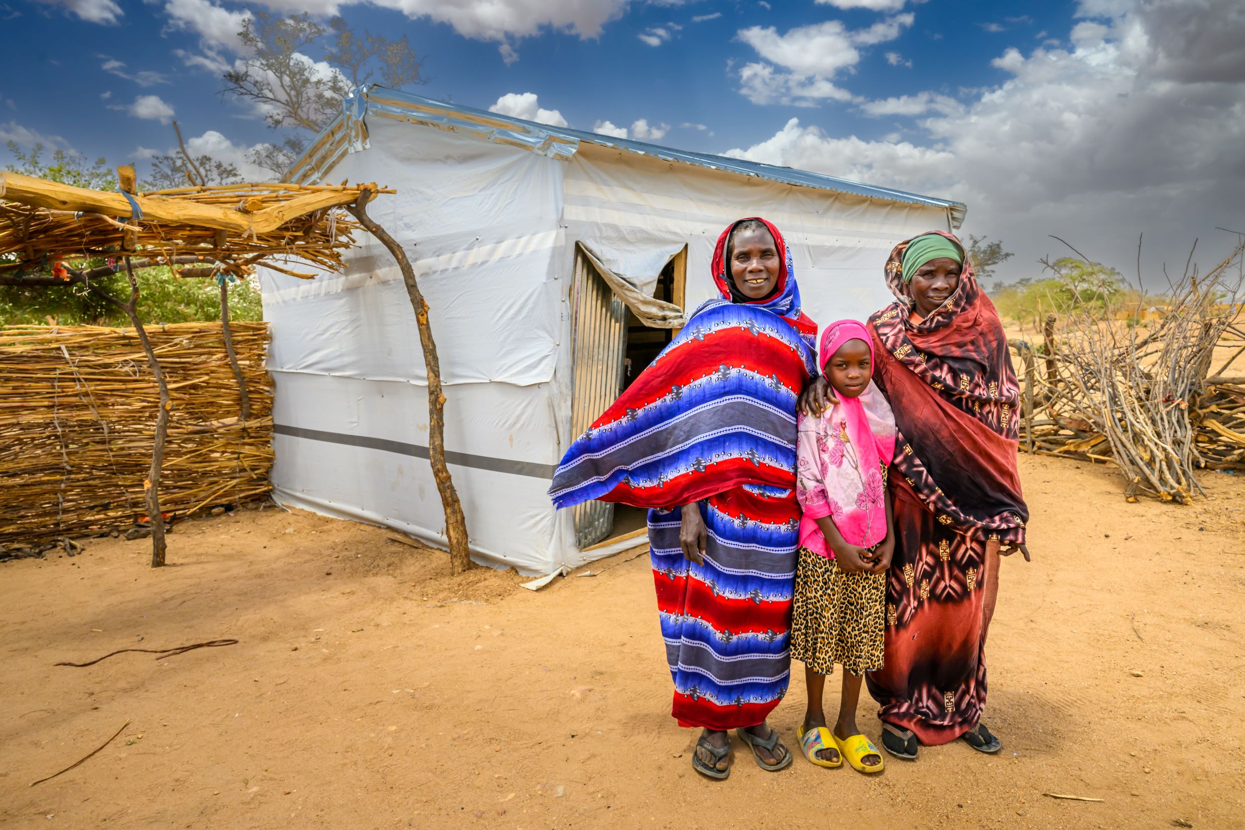 Sudanese girl stands between her aunt and grandmother outside their tent in a displacement camp, Chad