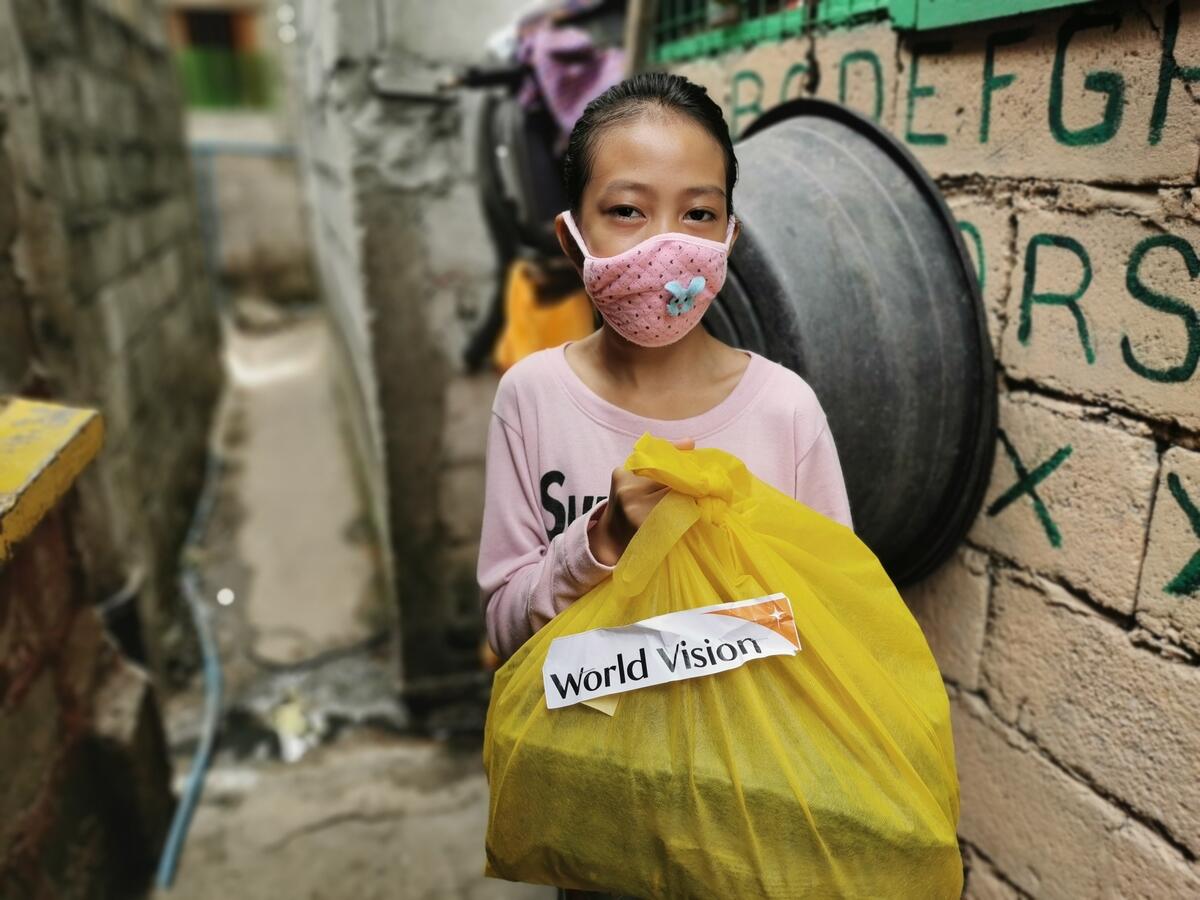 A young girl from the Philippines stands outside her house. She's wearing a mask to protect herself from coronavirus and holding a yellow bag of supplies with a World Vision logo on.