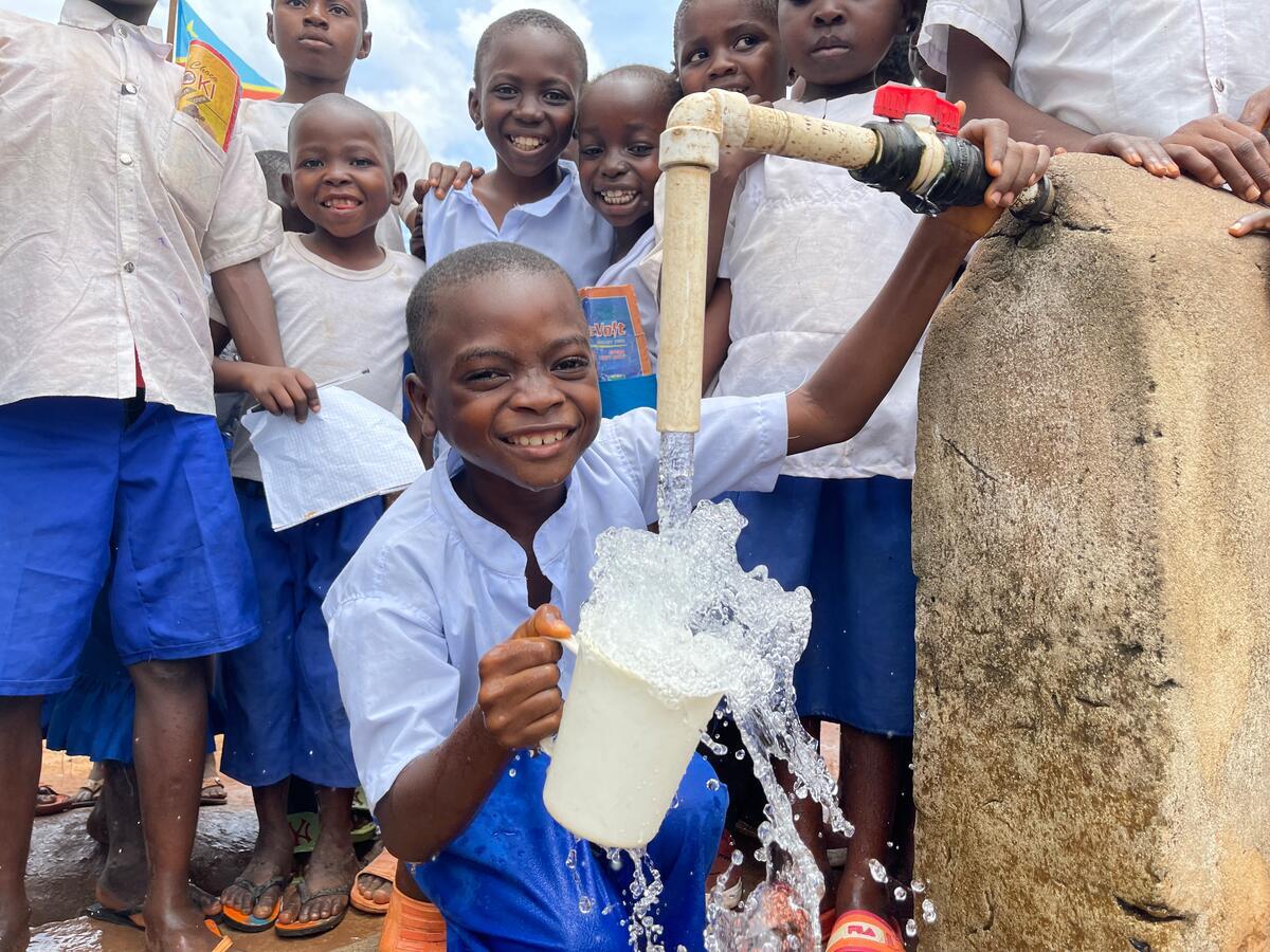 A young boy drinks clean water from a pump, with other children standing behind him