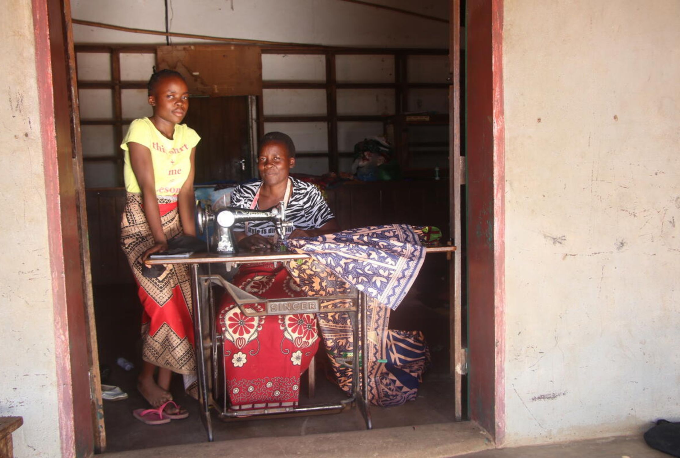 Zambian mother sitting and daughter standing with a sewing machine and smiling