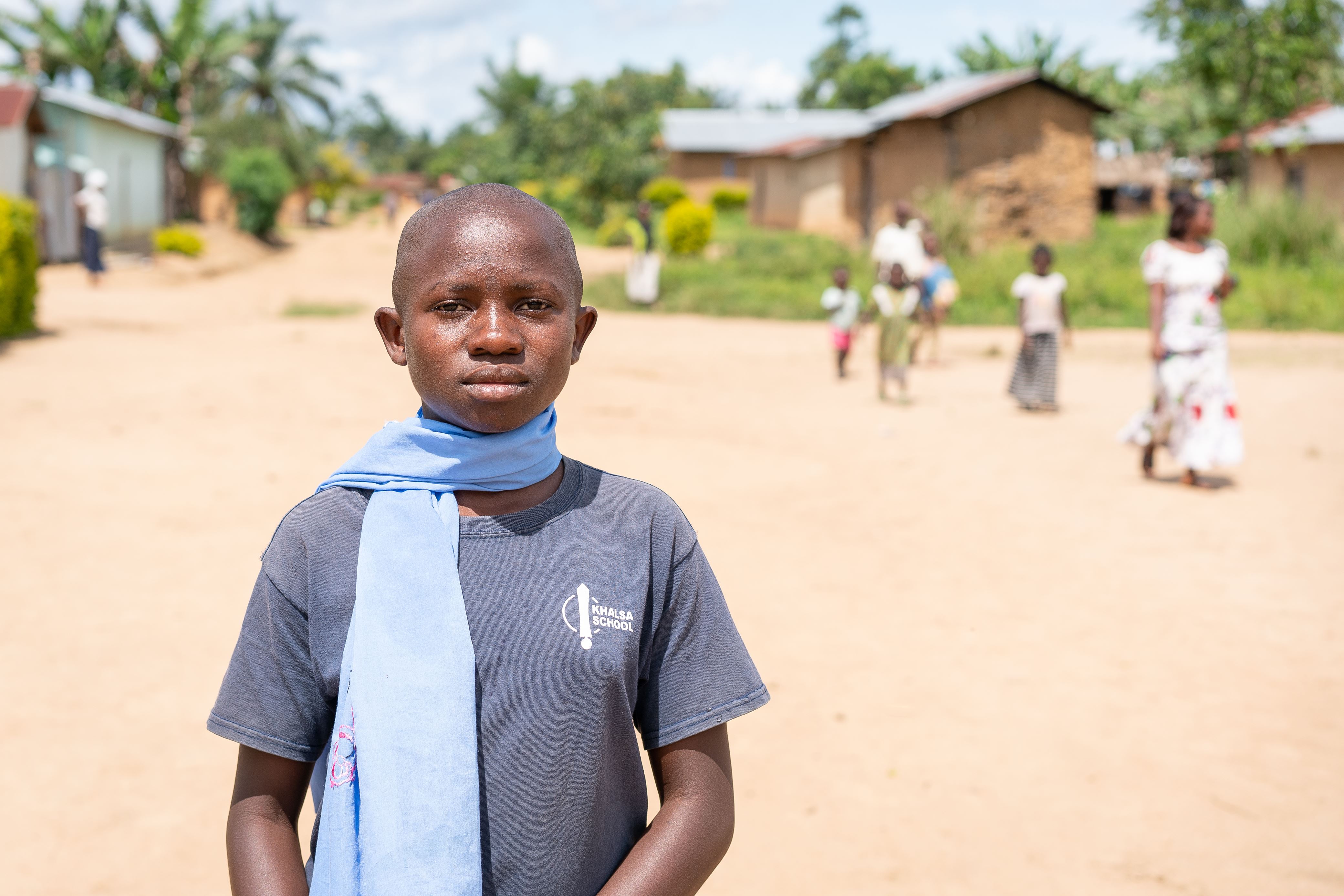 Girl in DRC stands and looks into the camera, wearing a blue top and blue scarf, standing outside with a few people walking around behind her
