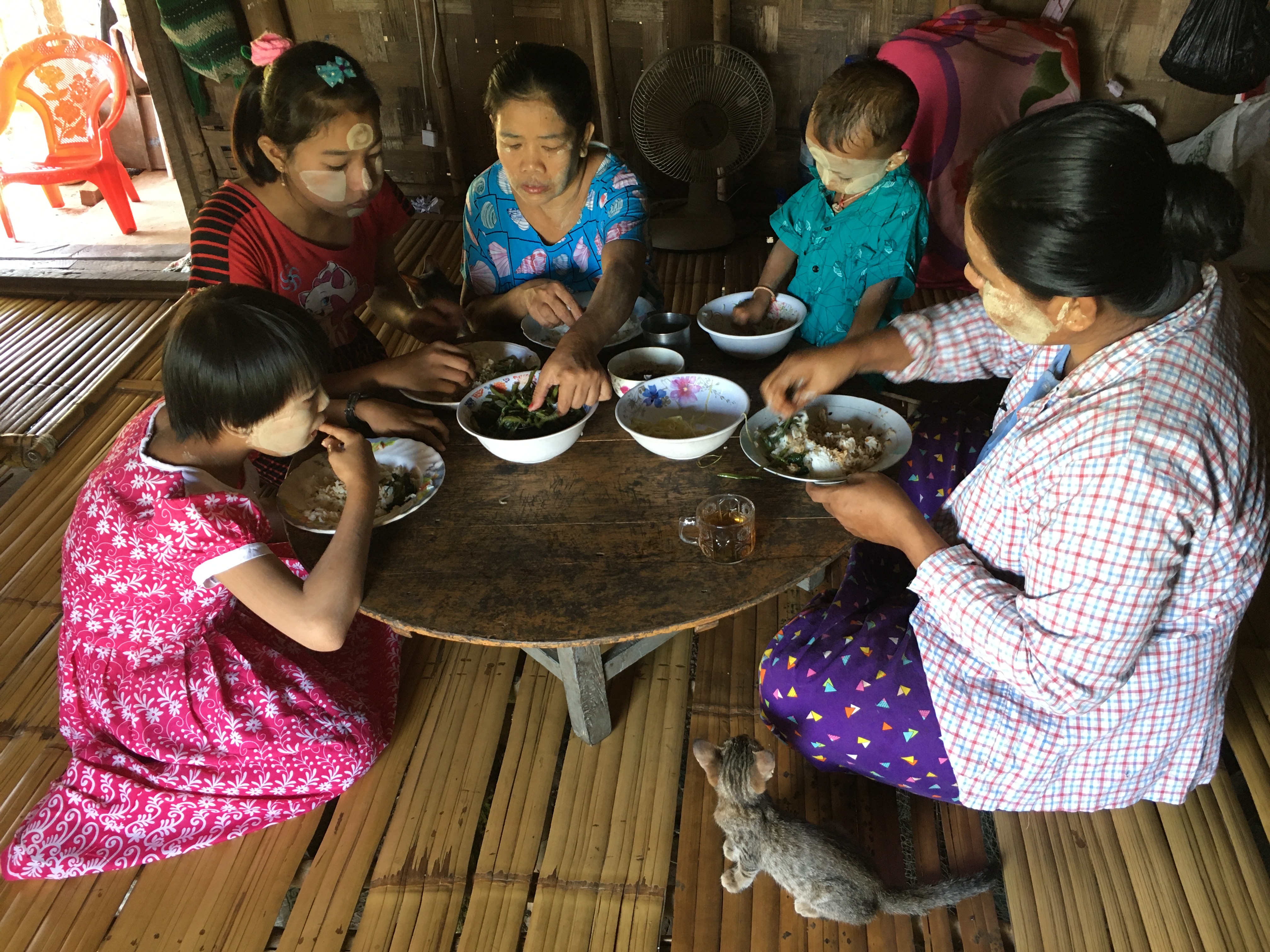 A woman from Myanmar and four children sitting in a circle on the floor around their dinner table and eating with their hands