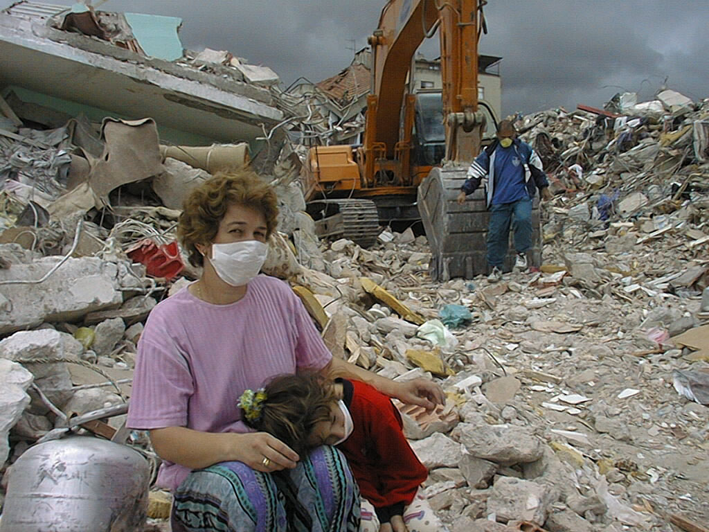 Woman and child wearing masks are surrounded by building rubble and digger searching for people under rubble