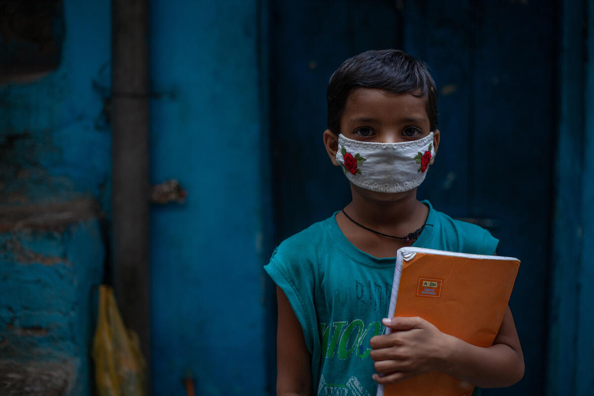 A young child in India stands in front of a blue-painted wall, wearing a face mask to protect against coronavirus