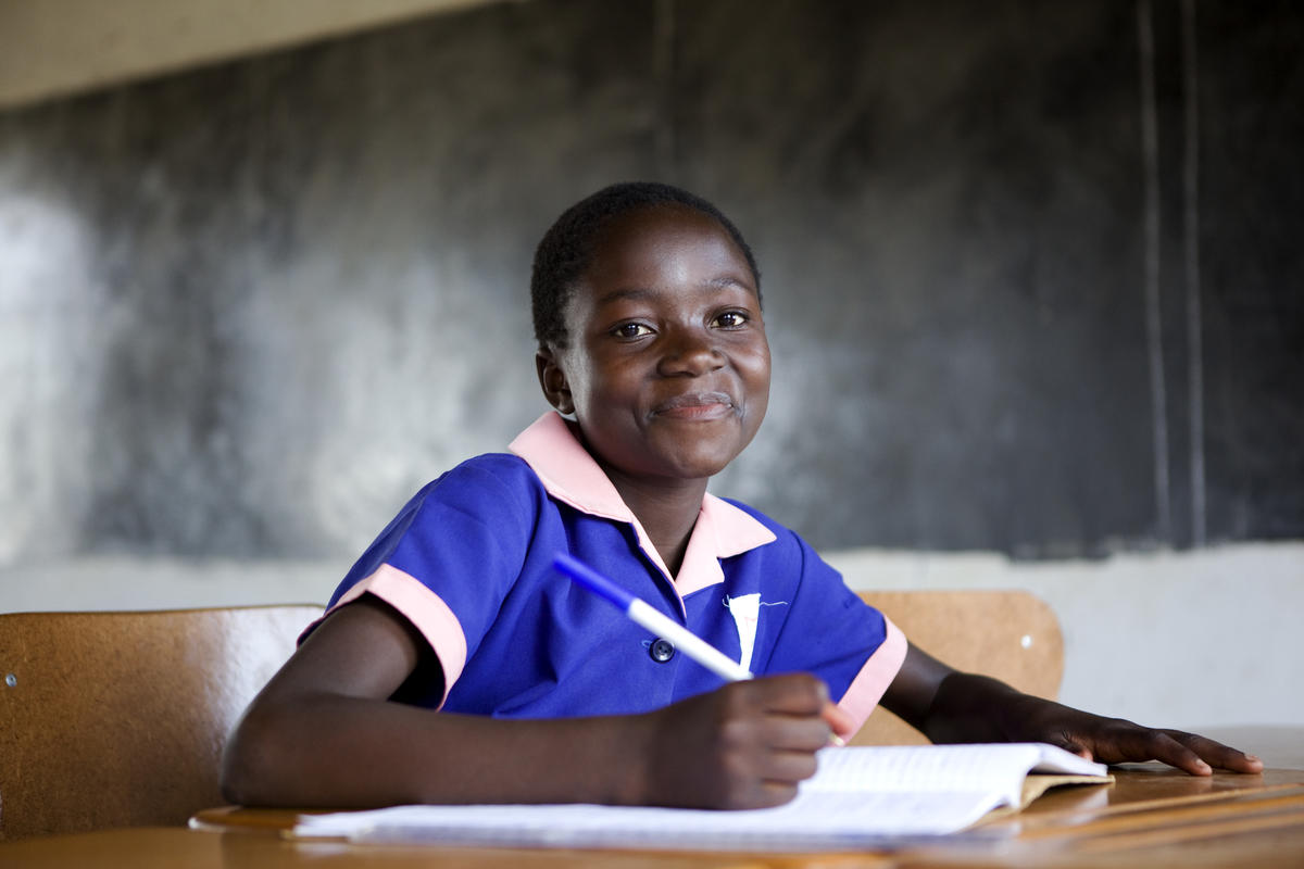 A teenage girl sits at her desk in a classroom, pen in hand, ready to work and smiling at the camera