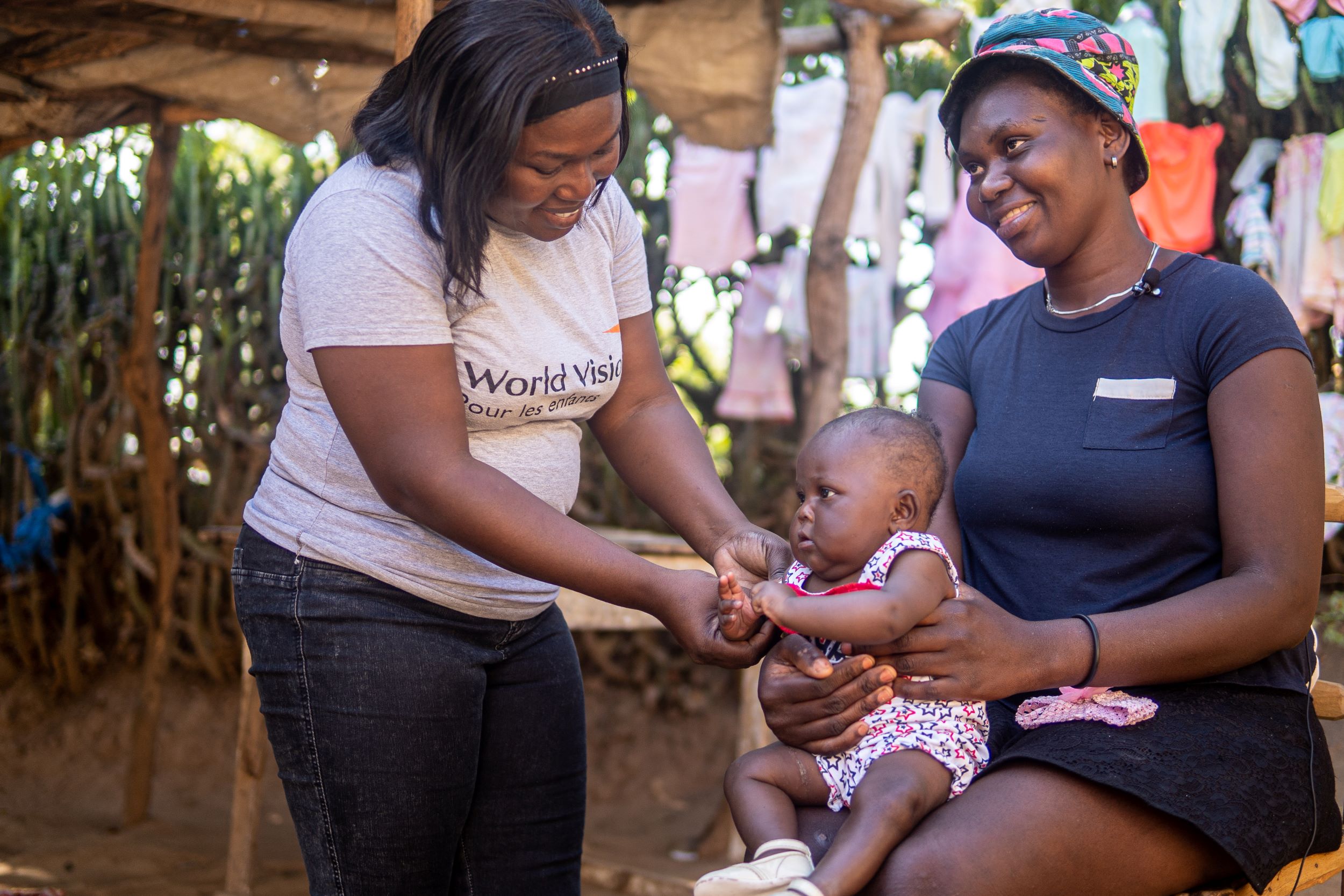 A woman sits, smiling and holding her baby while a World Vision staff member checks her baby's health