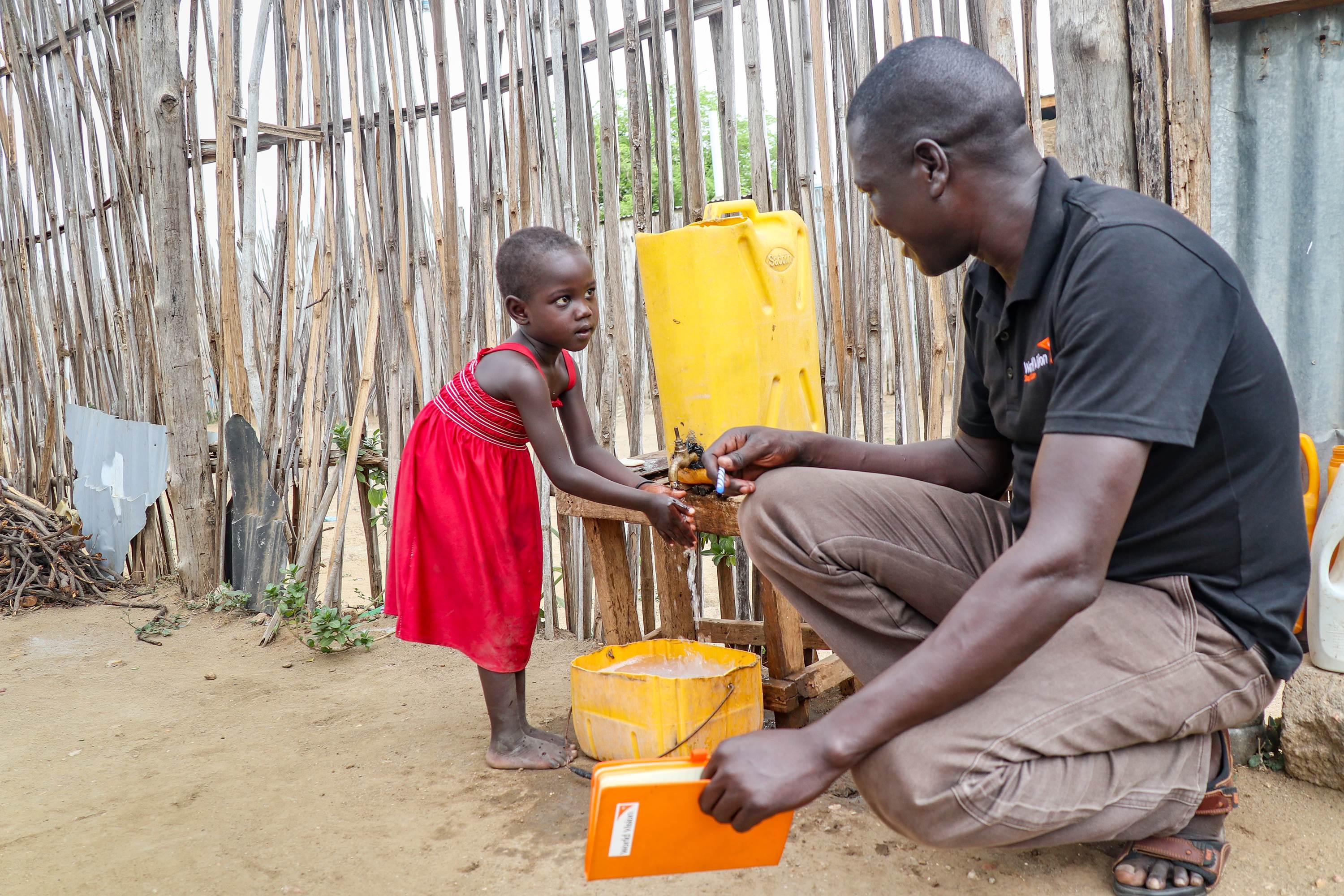Girl in a red dress in South Sudan smiles as she washes her hands in water from a yellow bucket, next to a man