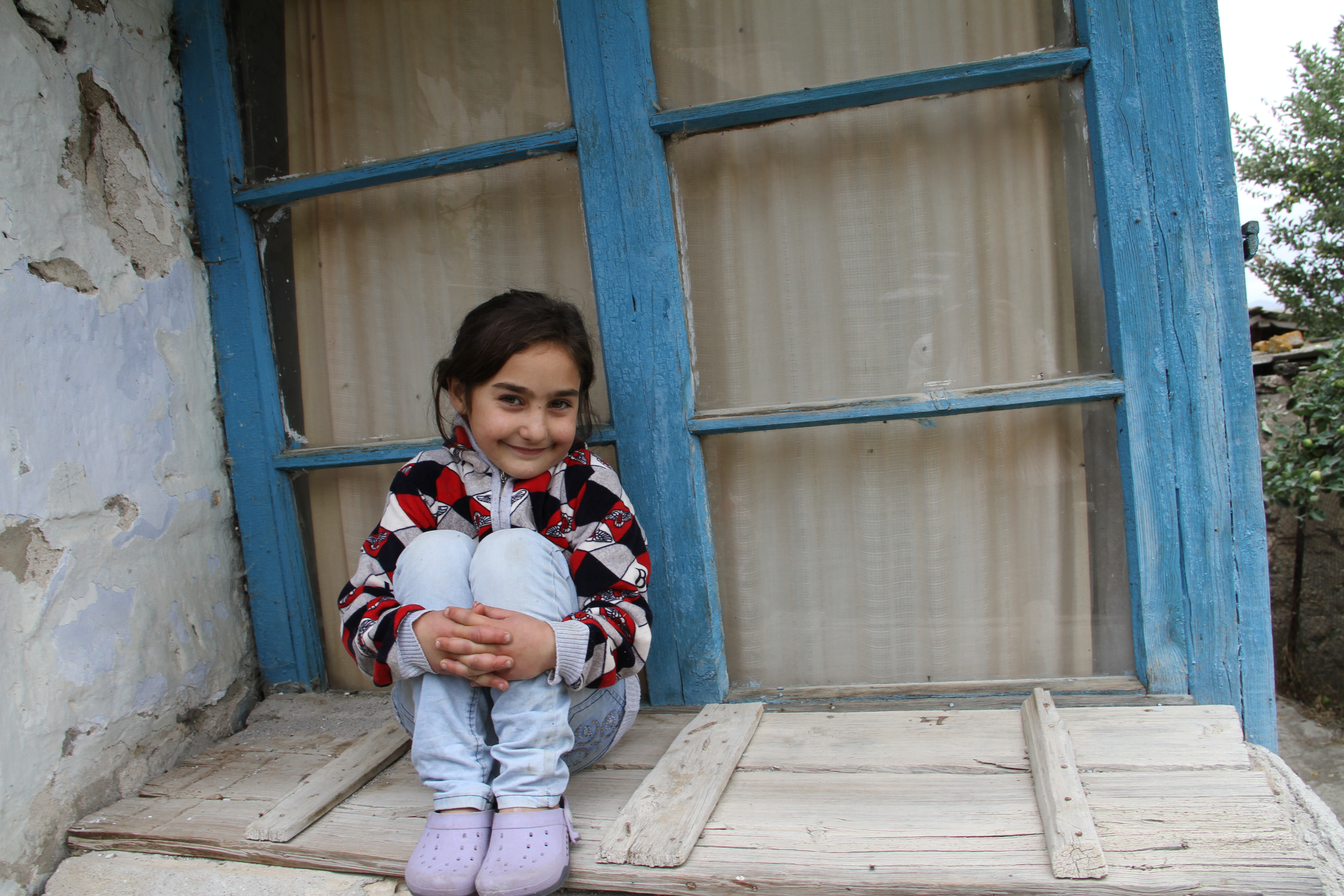 Girl sits with her arms around her knees, against the wall of her house in Armenia