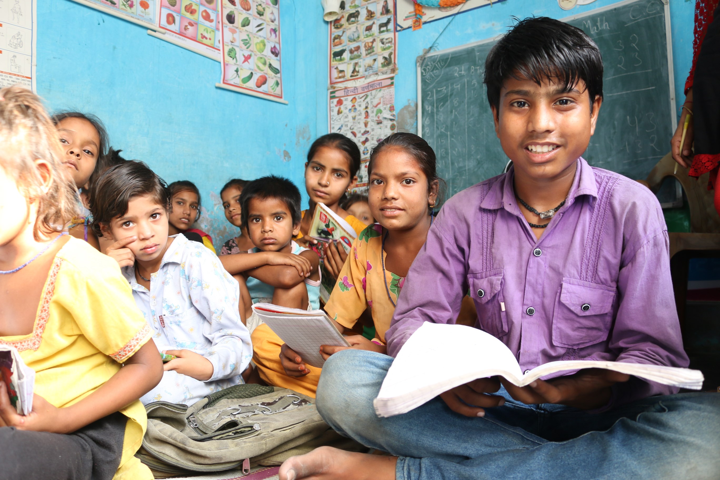 Boy sits in a classroom in India, cross legged and holding a book, surrounded by his classmates
