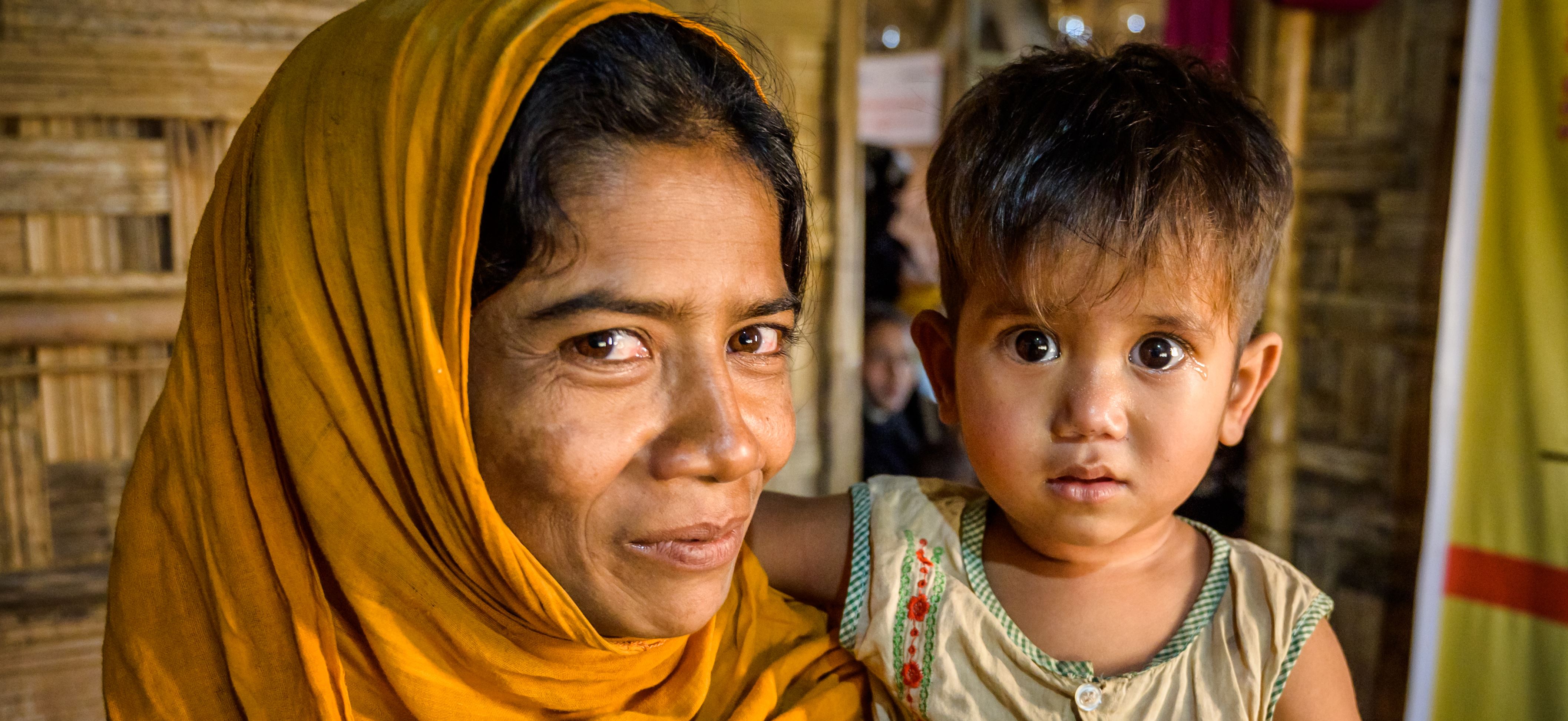 2 year old girl is held by her mother, who both look into the camera. She suffered from malnutrition when they first arrived in Bangladesh as refugees but now is a healthy weight.