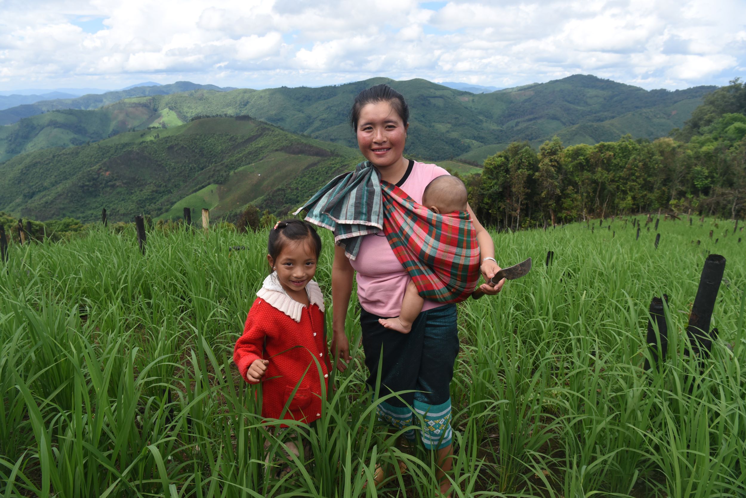A mother poses with her daughter and baby in a sling among tall green crops