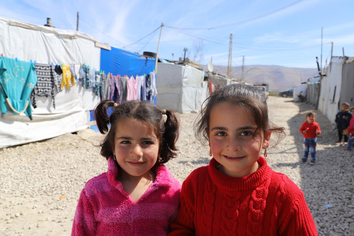 2 Syrian children smile, standing outside in front of tents in a refugee camp in Lebanon