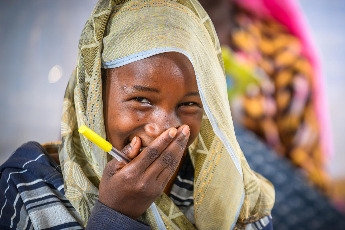 A young girl smiles with her hand covering her mouth, she holds a yellow pen and is laughing at the camera 