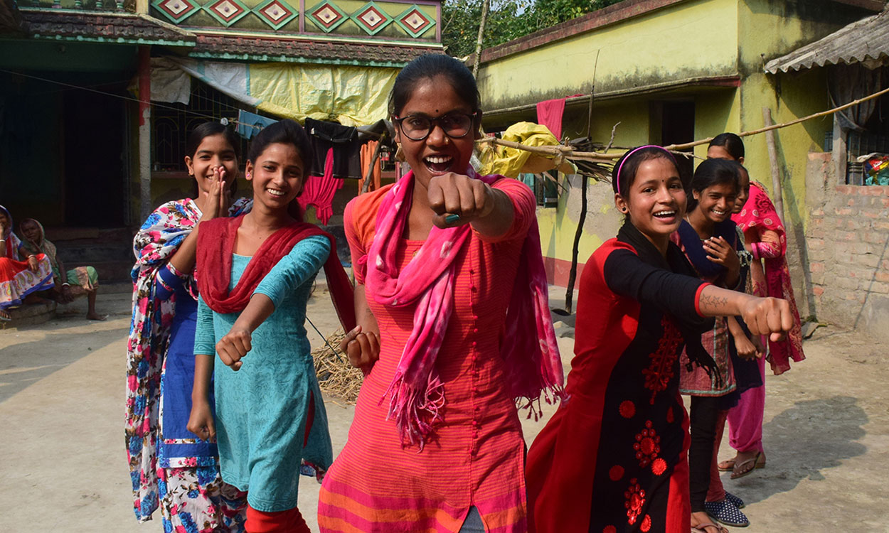 A teenage girl from India laughs at the camera while a larger group of girls stand smiling behind her