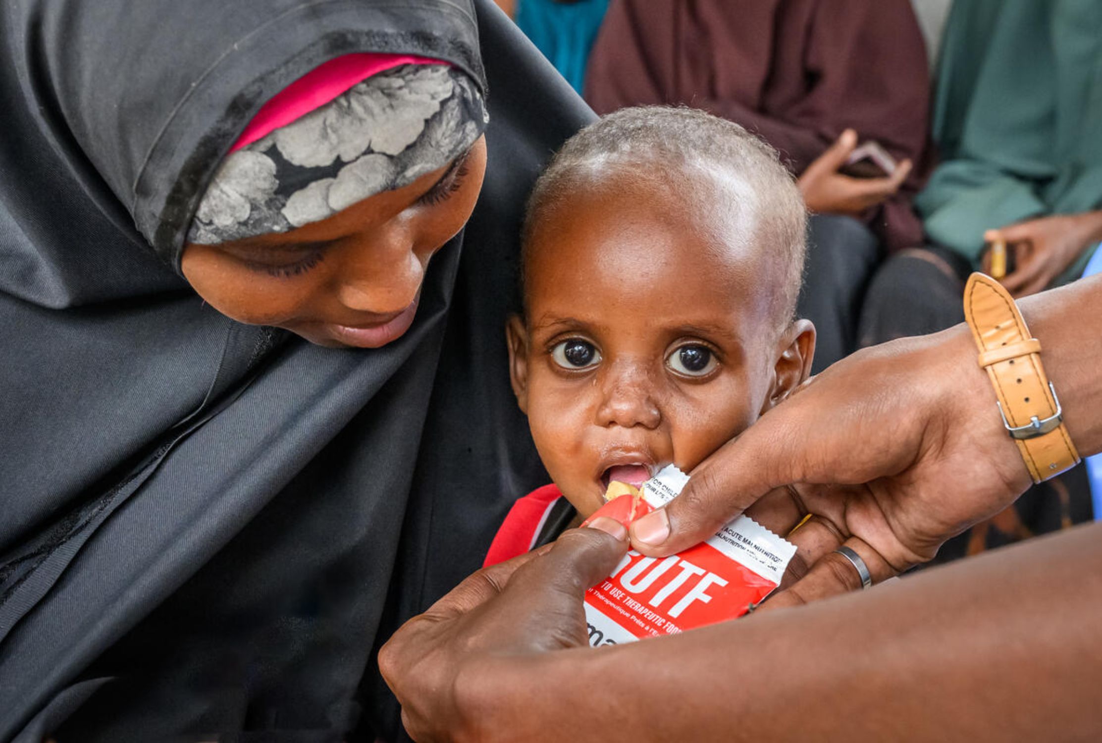 A mother from Somalia, holds her child in her arms, while another person helps them eat from a packet of therapeutic food.