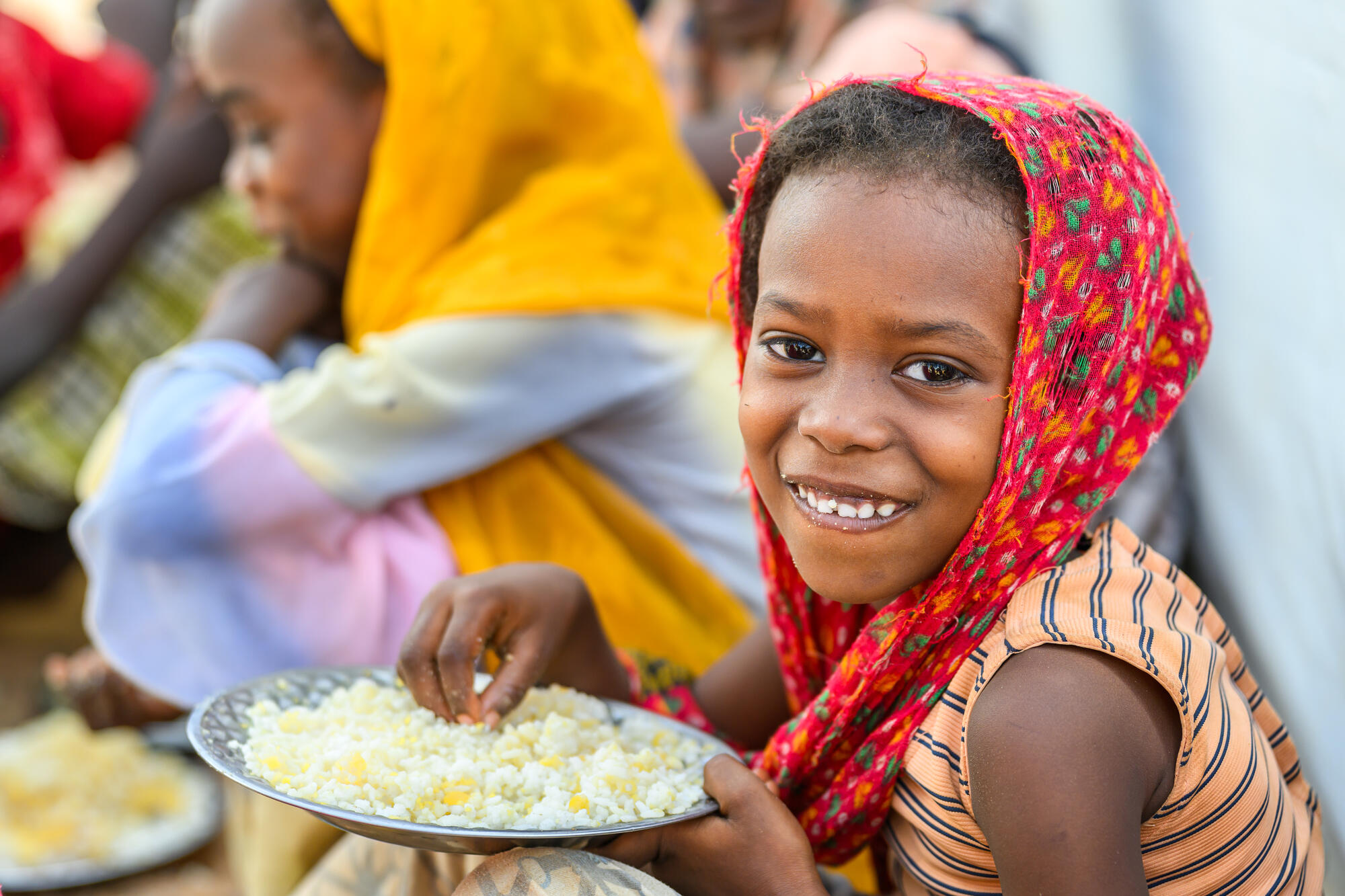 A young Sudanese girl smiles at the camera with a plate of food 