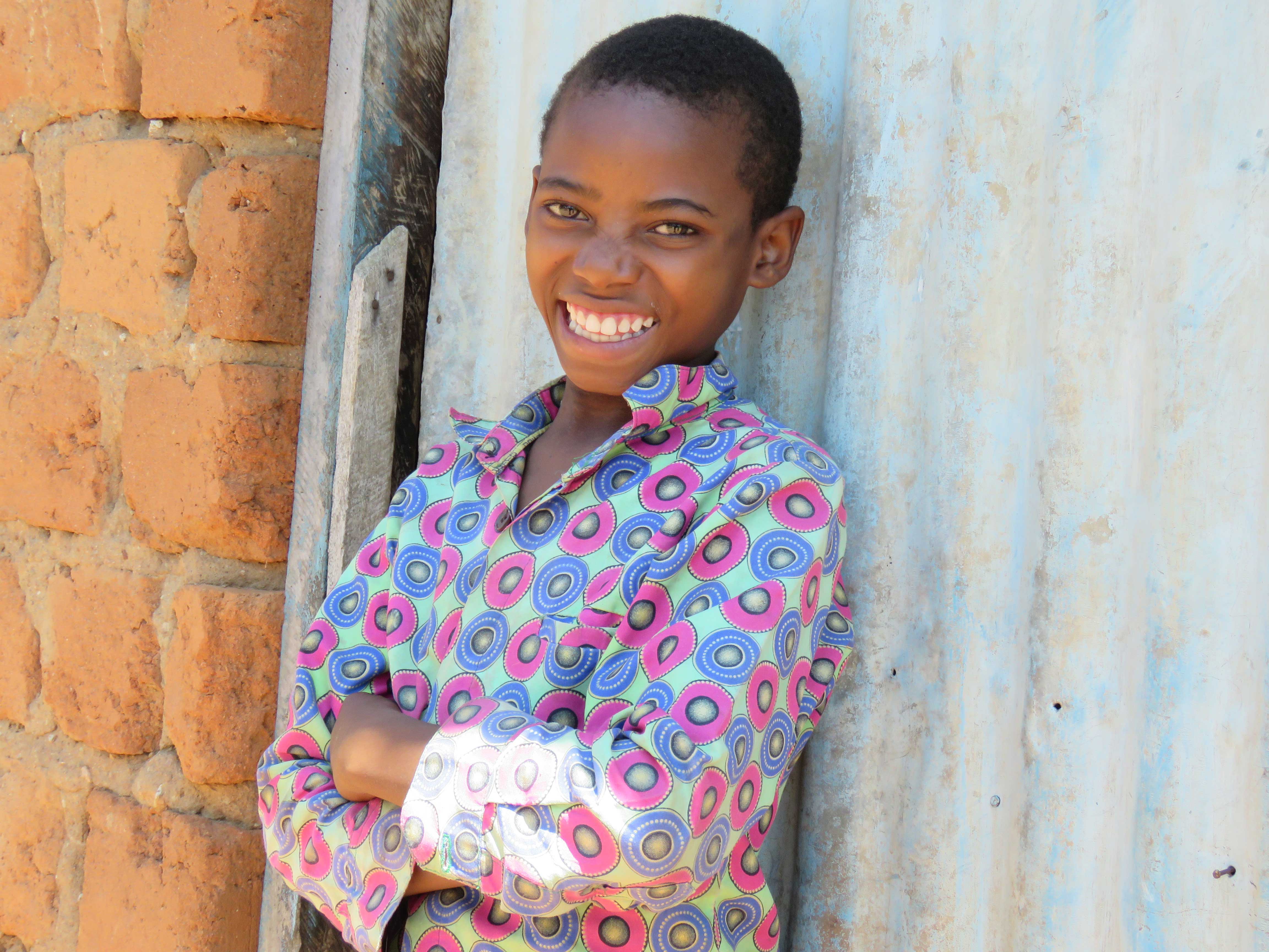 Boy from Tanzania smiles broadly as she stands next to a wall