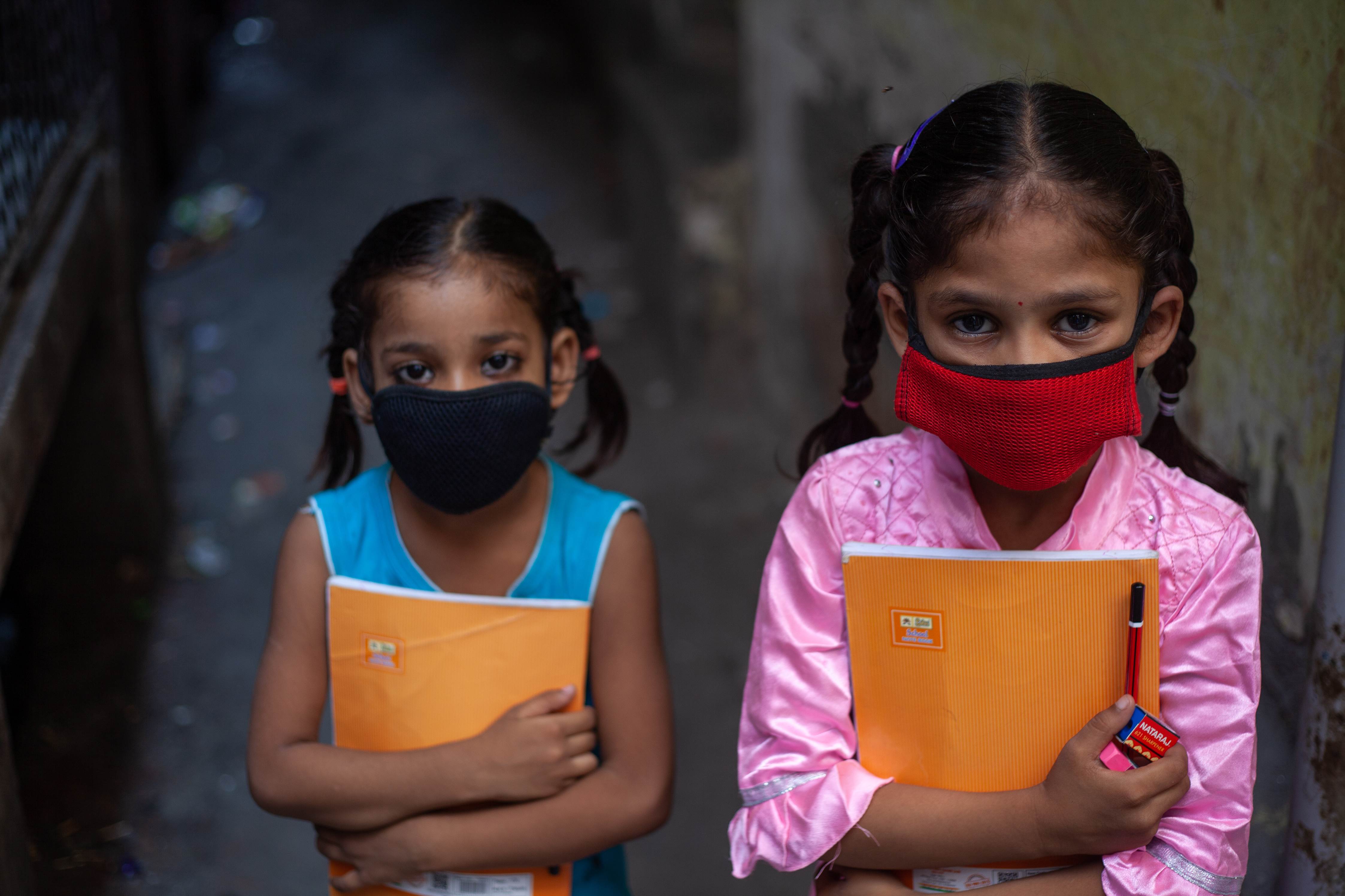 Two young girls, with plaits, wear face masks and hold their school books tightly