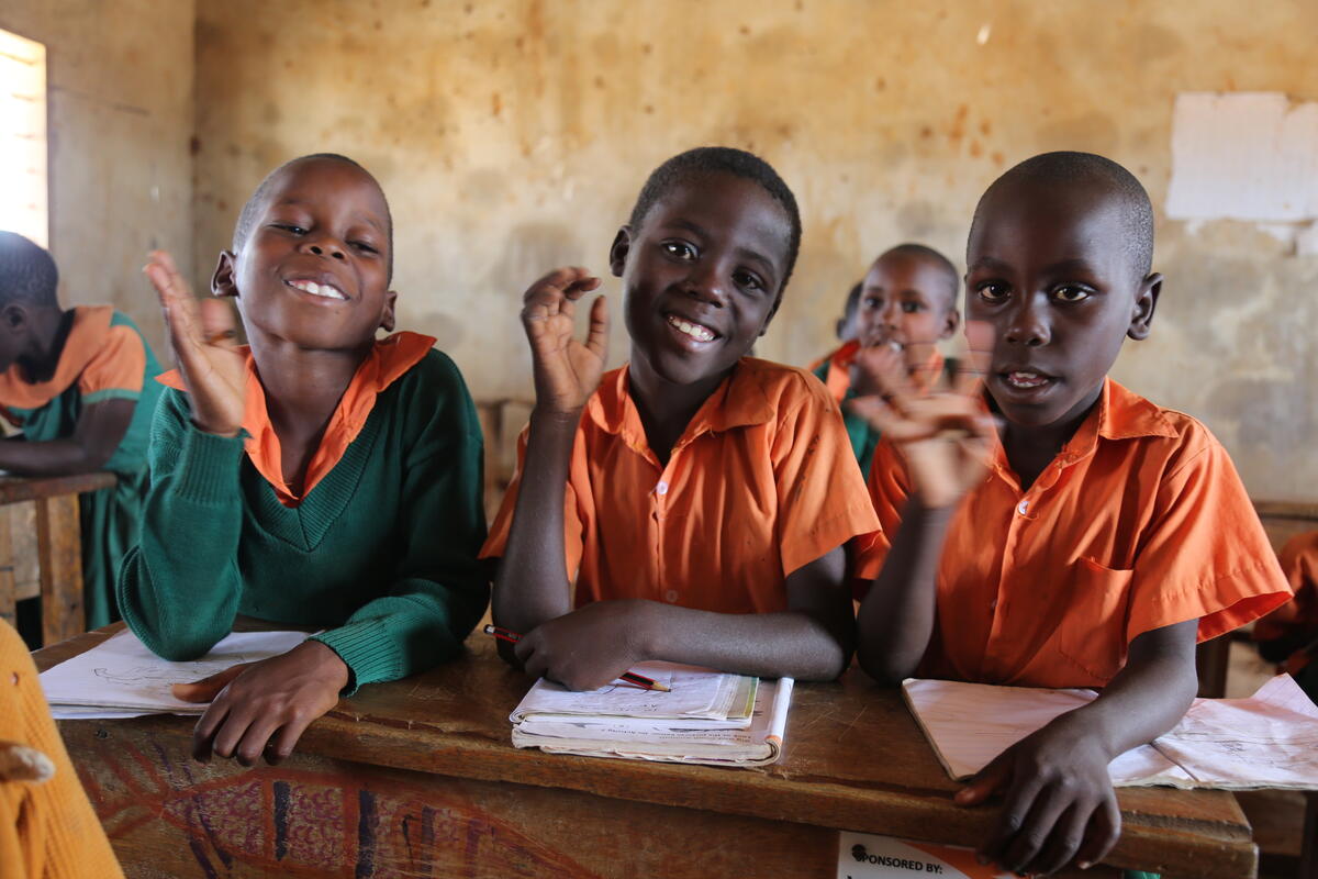 Three Kenyan school children sat at a desk, waving and smiling at the camera