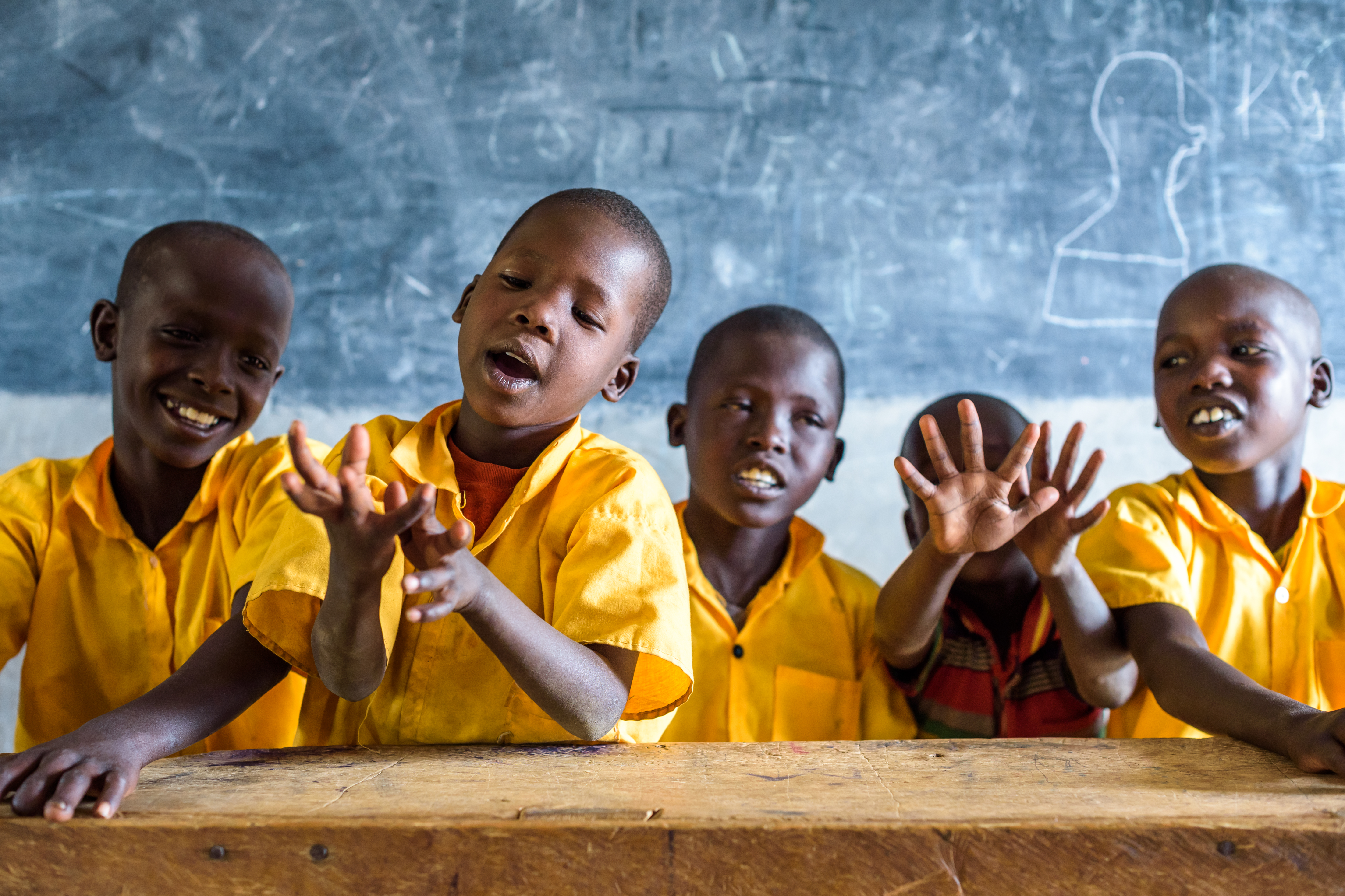 Four children from Kenya smile and chat as they sit in a classroom, in front of a blackboard, all wearing bright yellow tops