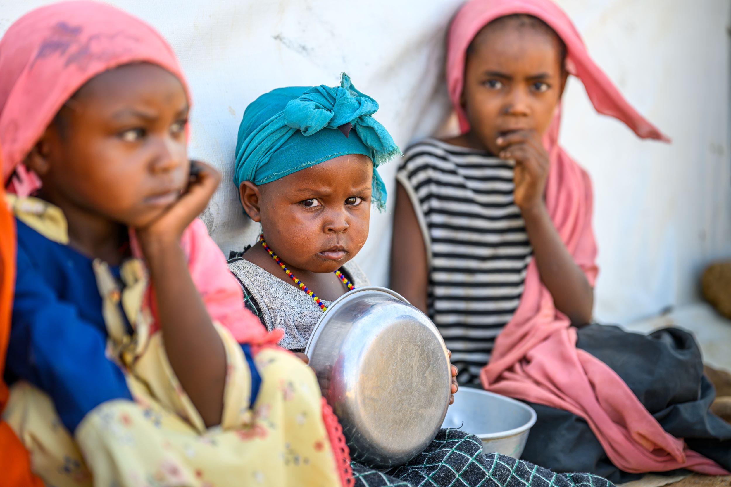 Three young girls from Sudan sit on the floor holding metal food bowls