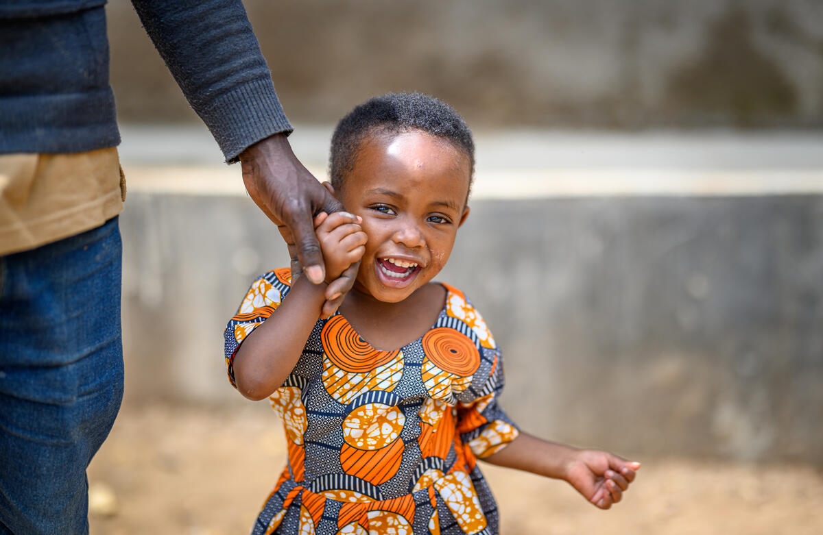 A young girl in Rwanda smiles as she clings to the hand of an adult