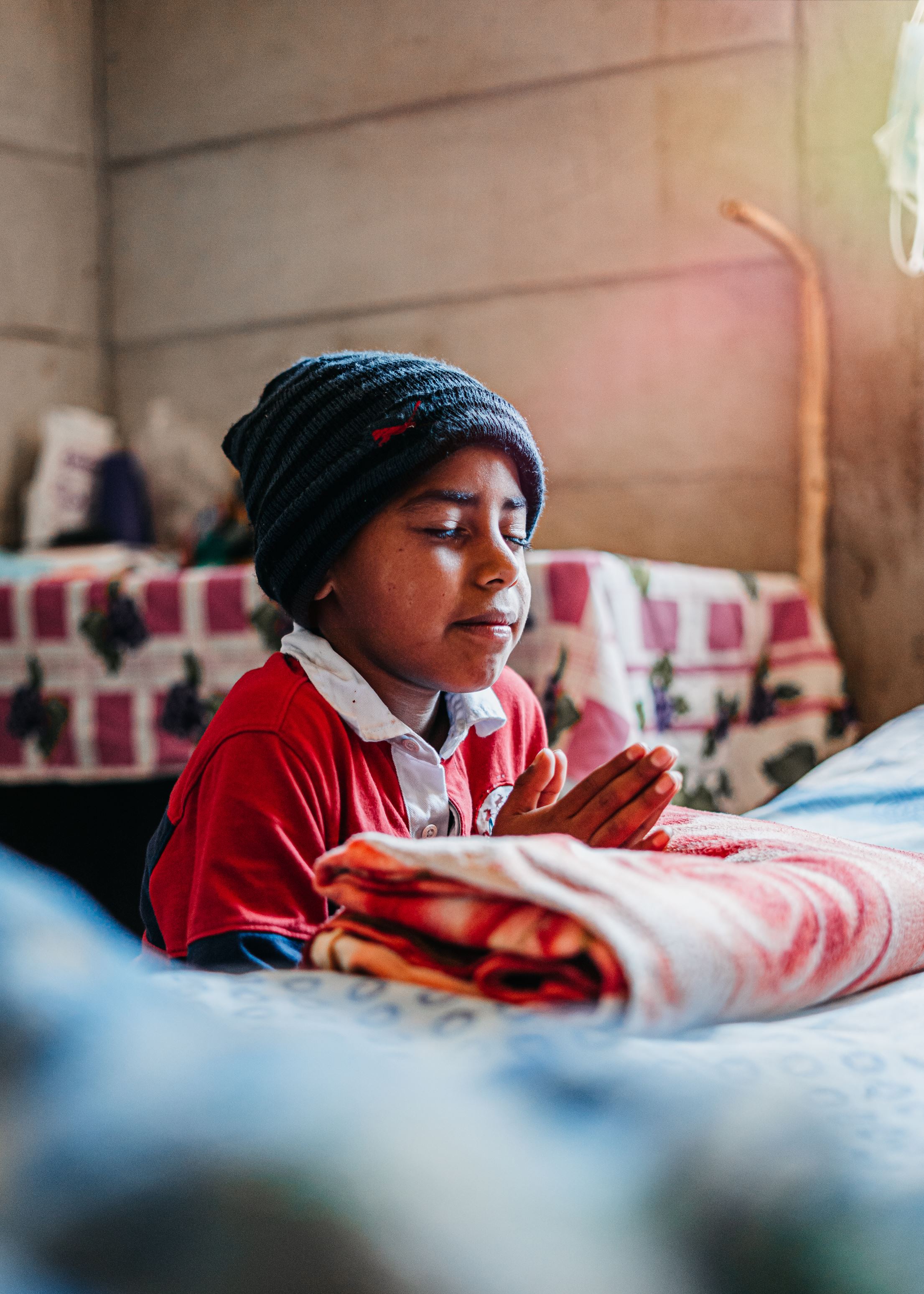 Young boy in red with a hat on knelling on the floor by his bed praying with his hands out in front of him.