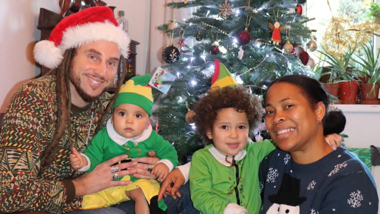 Family of 4 in the UK smile as they sit in front of a Christmas tree, a baby dressed up as an elf