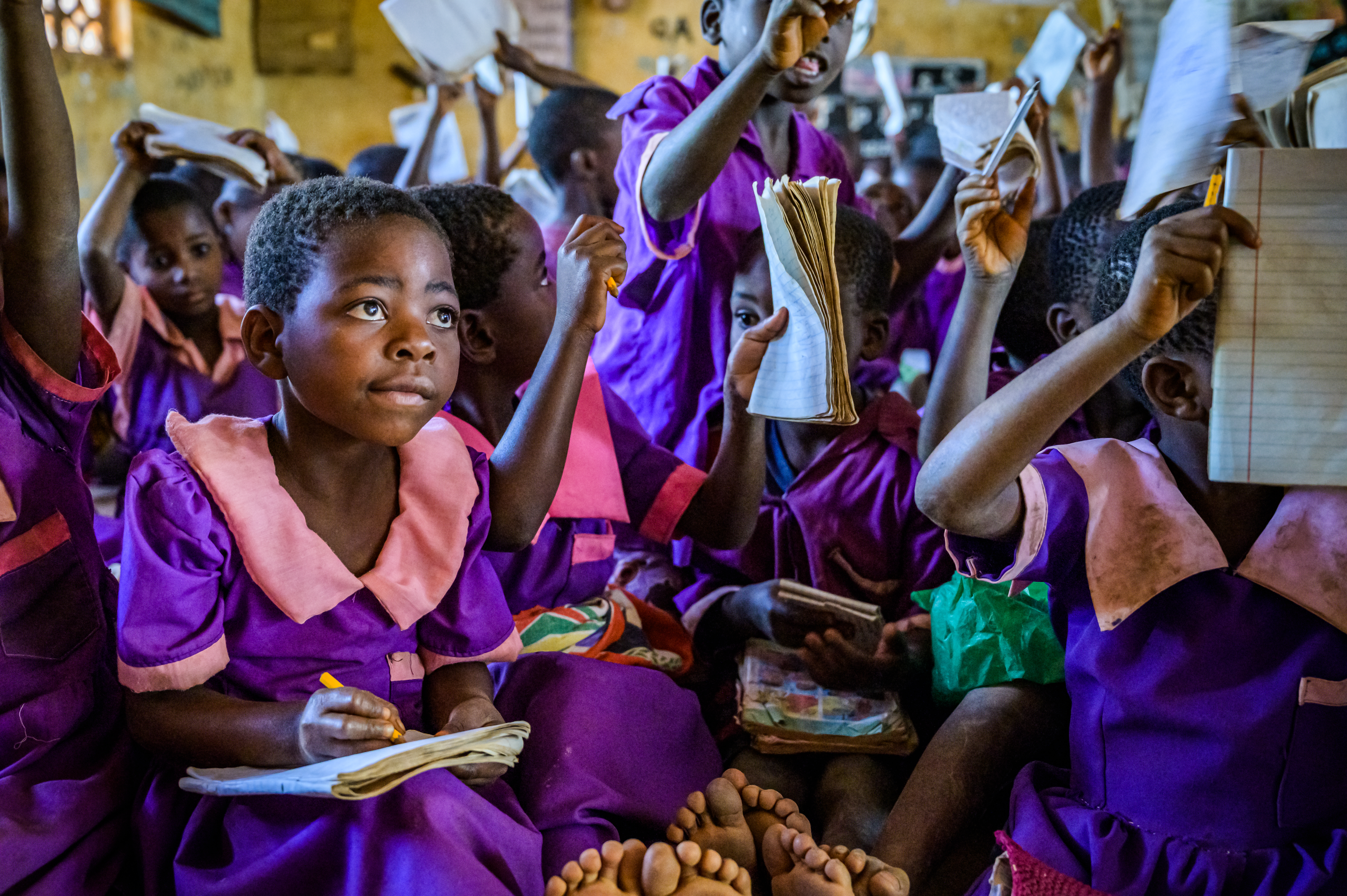 Girl in Malawi sits in a classroom with her peers, many with arms raised to answer a question