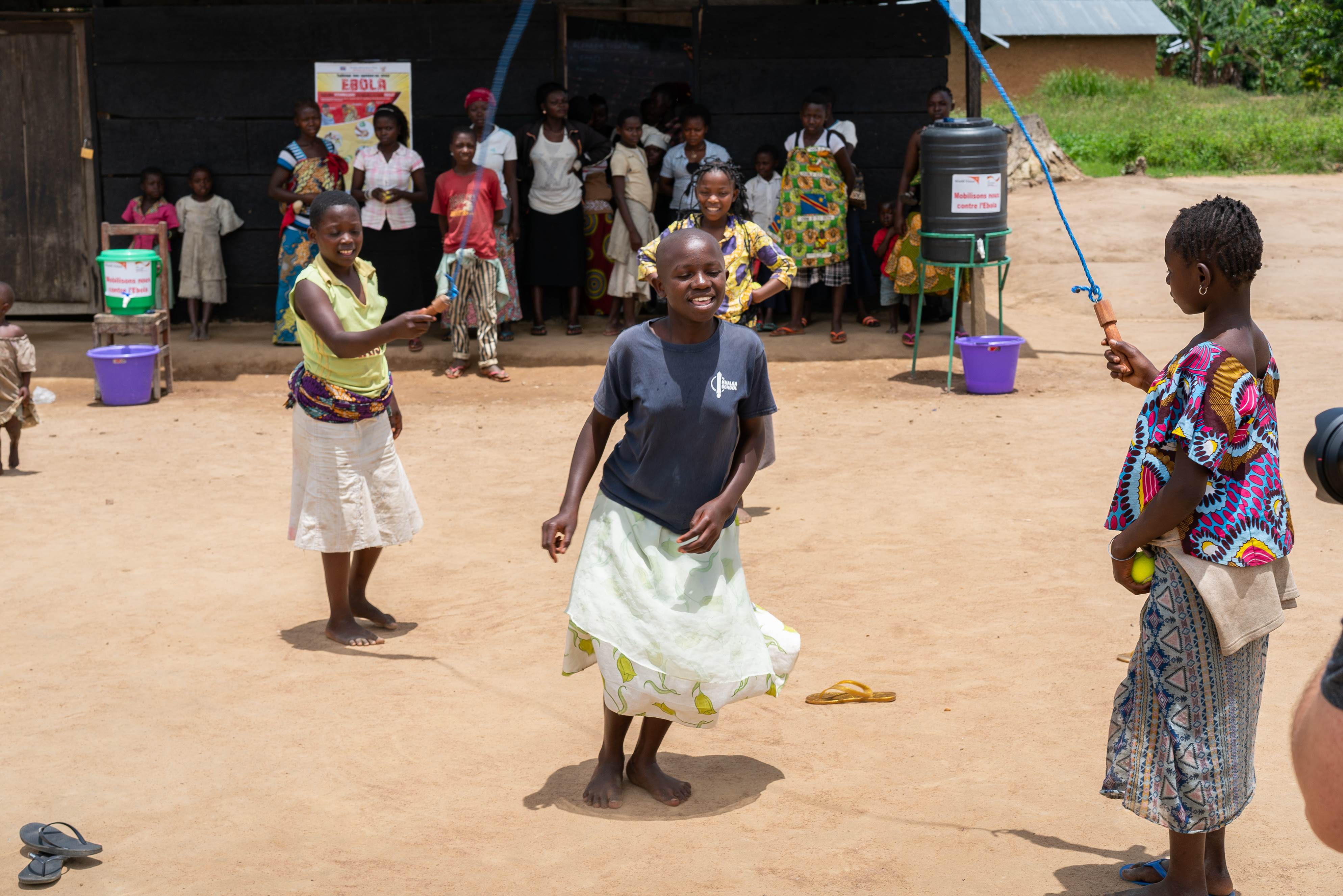 Girl in DRC skips over a rope, wearing a blue top and blue scarf and yellow skirt, outside with a few people behind her watching, and two others holding the rope