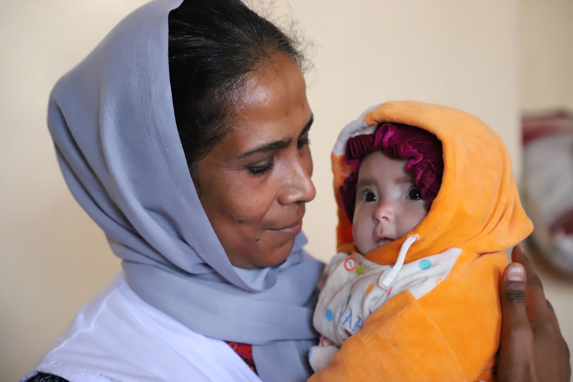 A woman, with blue headscarf, holds a toddler, wearing yellow