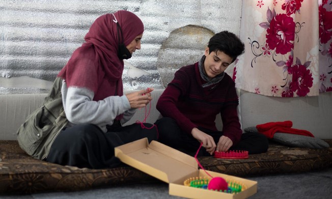 15-year-old Syrian refugee, Muath, sits next to his mother, practicing knitting and crochet, in Azraq refugee camp
