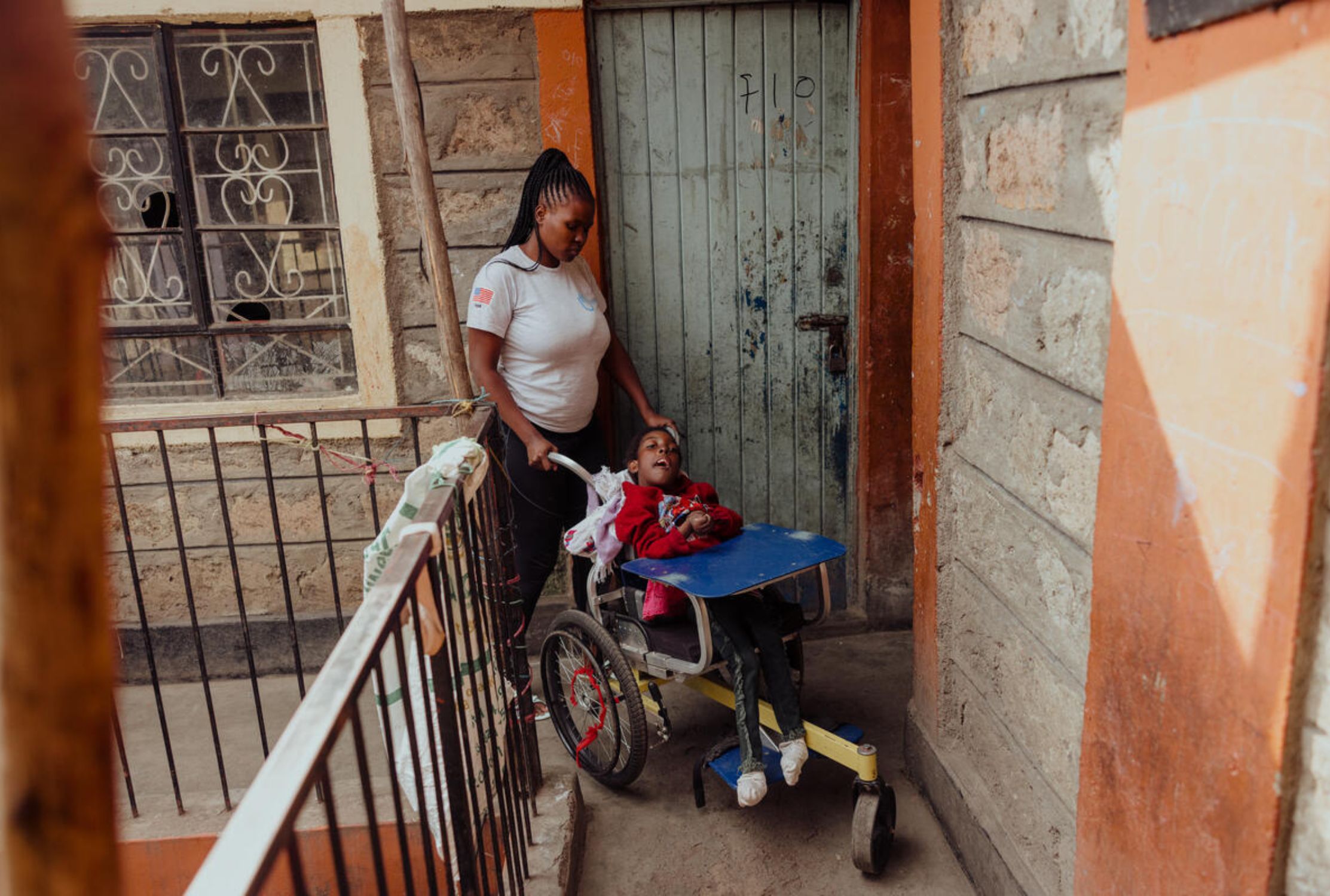A girl with cerebral palsy sits in a wheelchair while her older sister pushes her down the corridor of their building